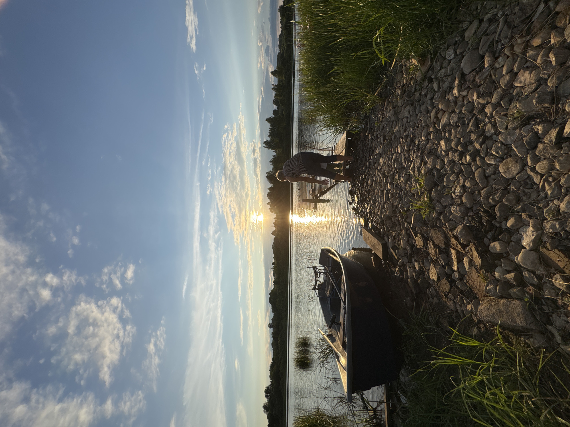 At sunset on a body of water in Sitno, Russia, a person is standing on a wooden pier, facing away from the camera. The person is wearing a striped shirt and dark pants. A small boat is moored to the pier, and tall grass grows along the water's edge. The sky is blue with scattered clouds, and the sun is setting on the horizon, casting a golden glow on the water. The shoreline is covered in stones and dirt.