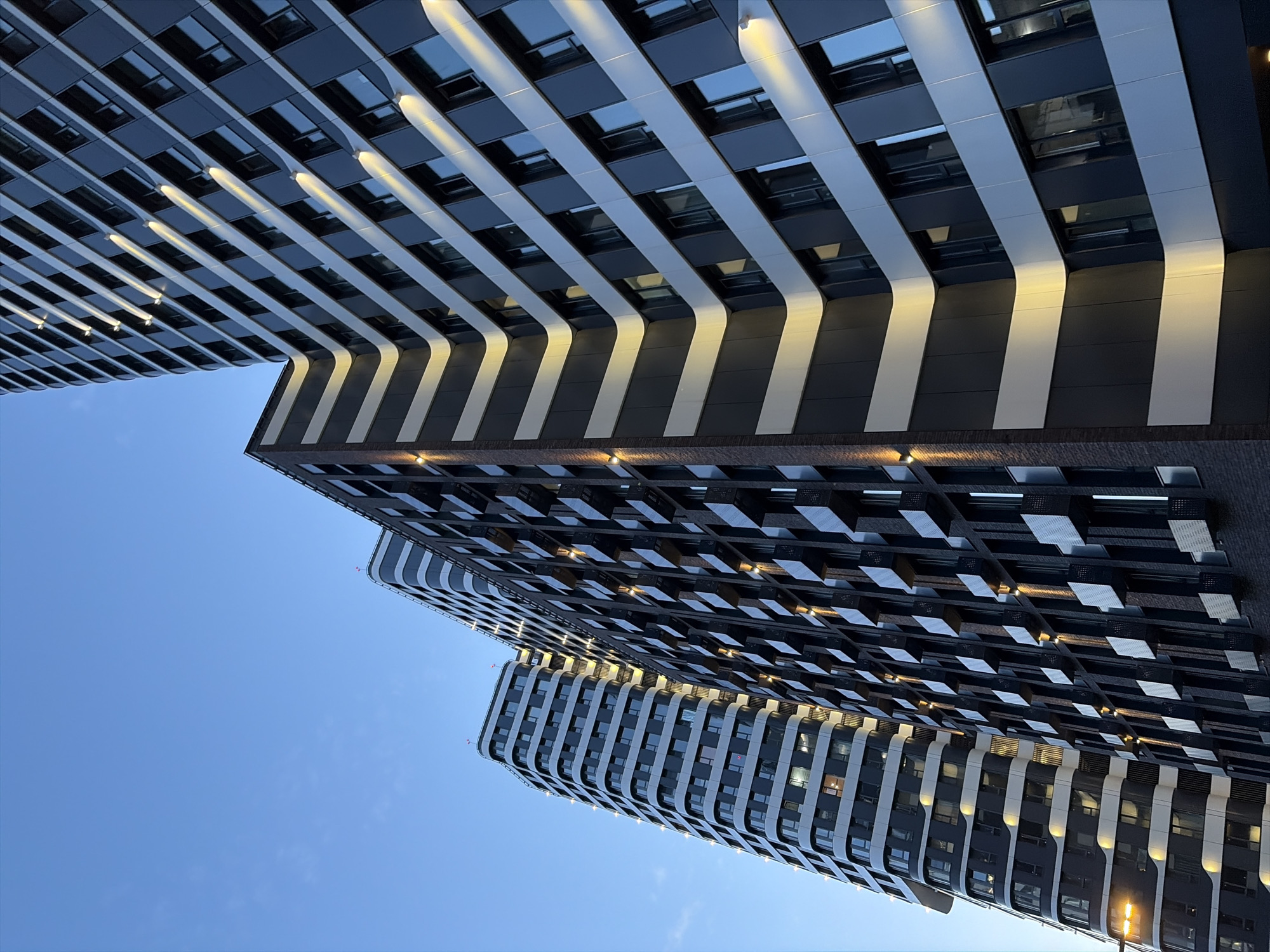 The image captures a section of a modern residential complex in Moscow, Russia, with a clear blue sky as the backdrop. The architecture features multiple high-rise buildings with distinct designs. One building on the left has a curved facade with horizontal bands of white and dark gray, punctuated by numerous windows. To its right, a taller building with a more angular design displays a repeating pattern of dark gray panels and recessed window openings, many of which appear to be balconies with small enclosures. Further to the right, another building with a similar dark facade and window pattern rises. The buildings are illuminated by a warm, yellowish light that highlights the architectural features, suggesting it is either dawn or dusk. There are no people or vehicles visible in the frame. The overall scene conveys a sense of urban development and contemporary residential architecture.