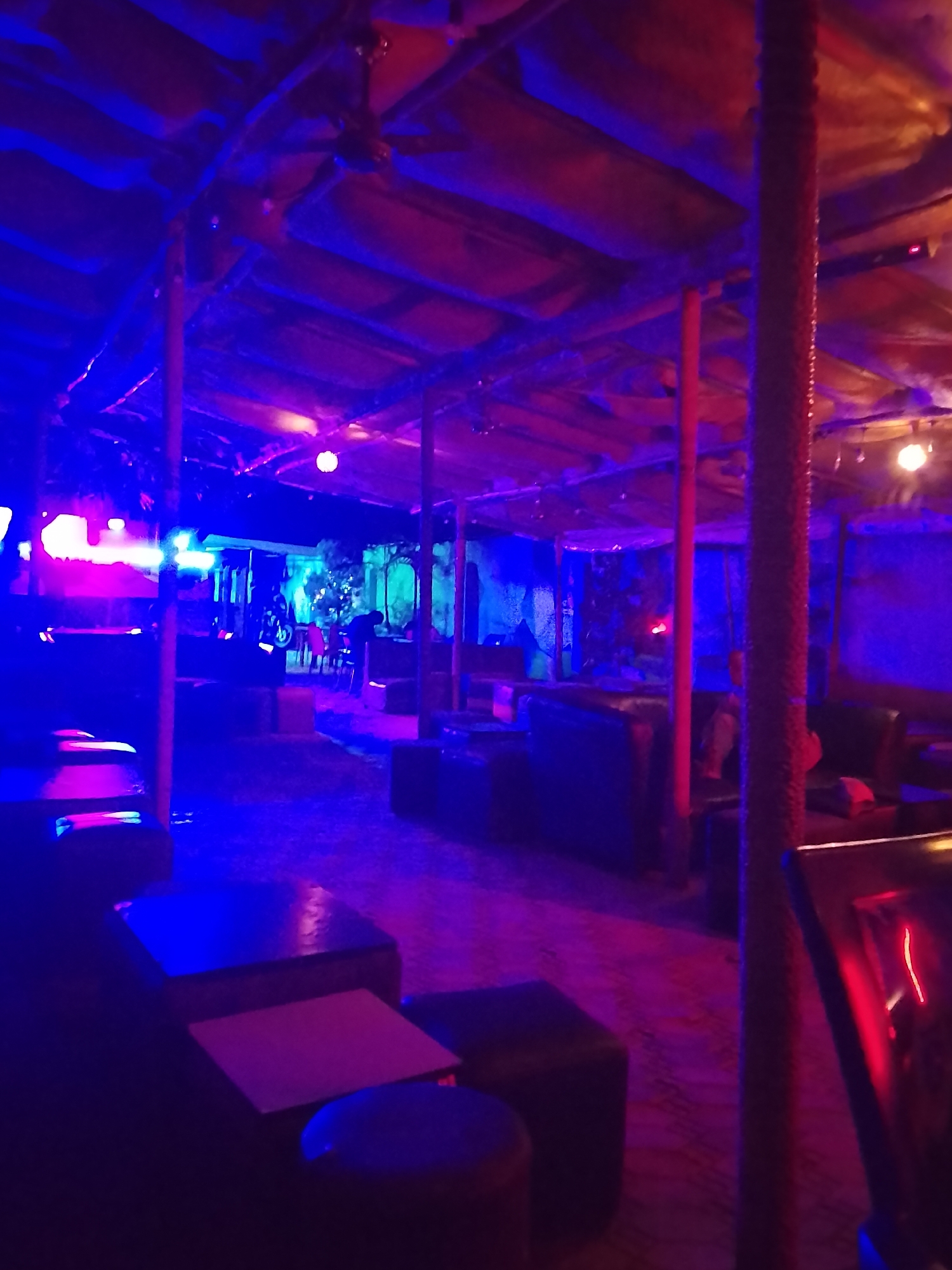 The image depicts the interior of a bar in an unknown city. The space is lit with blue and purple lights. The ceiling is low and appears to be made of fabric stretched over a frame. Wooden support posts run through the space. There are tables and seating areas with couches and stools. The flooring is patterned. In the background, an open doorway leads to an outdoor area with plants and a few patrons. In the foreground, there are unoccupied tables and stools.