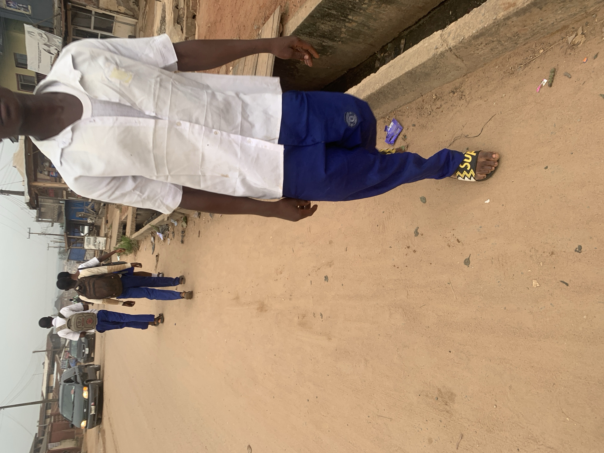The image depicts a street scene in Shagamu, Nigeria. In the foreground, a person wearing a white short-sleeved shirt over a grey t-shirt and blue trousers walks away from the camera. They are wearing yellow and black slide sandals. To the left of the foreground subject, three other individuals, likely students due to their matching uniforms, are also walking away from the camera. They are wearing white shirts, blue trousers, and backpacks. The individuals are walking on a dirt road, with a concrete drainage ditch to the right. Several vehicles, including a dark-colored minivan and other cars, are visible further down the road. The background consists of buildings and structures that suggest a bustling urban environment. The overall lighting suggests an overcast day.