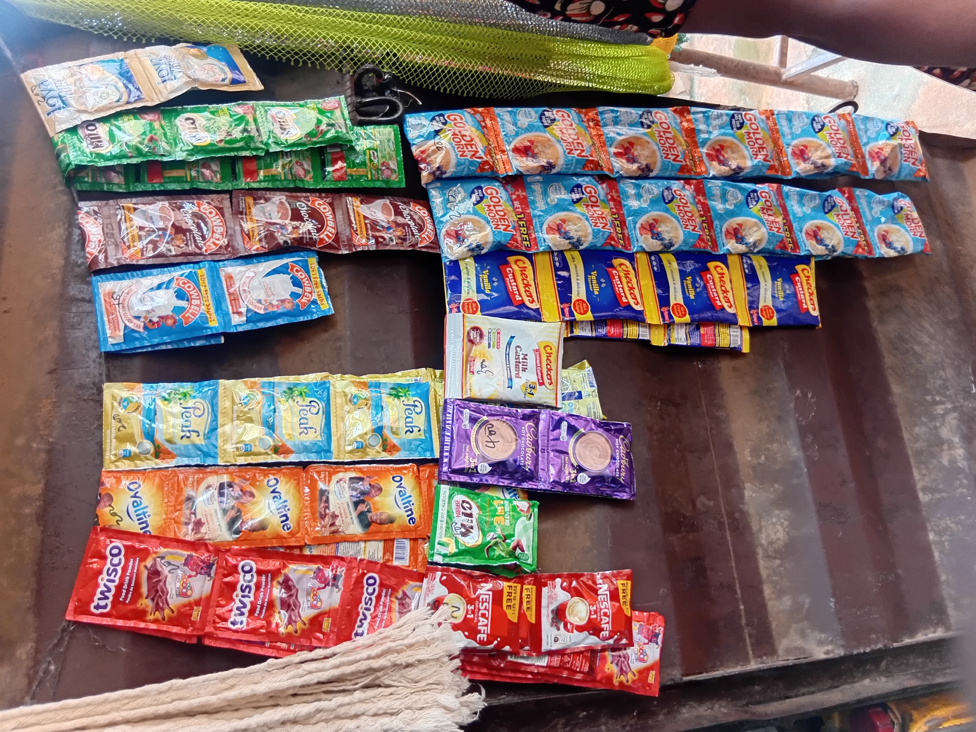 The image depicts a close-up view of a variety of food and drink sachets hanging from a display in an outdoor market stall, likely in Nkpor, Nigeria. The items are arranged in vertical rows and columns, secured with clips or hooks. Dominant brands visible include "Twisco," "Ovaltine," "Peak Milk," "Cowbell Chocolate," "Nestle Milo," "Cadbury Hot Chocolate," "Checkers Custard Powder," and "Golden Morn."
The products are predominantly packaged in colorful, foil-like sachets, featuring brand logos, product names, and imagery related to their contents, such as chocolate drinks, milk, and breakfast cereals. The background is a dark, weathered surface, possibly the corrugated metal wall of the stall, showing signs of rust and age. To the left, a bundle of thick, cream-colored rope or twine is visible, adding a textural element. To the right, a bright, lime-green mesh net hangs, and a portion of a person's arm and leg, wearing a patterned fabric, is visible, suggesting the presence of a vendor or customer. The lighting is somewhat dim, with the focus clearly on the displayed products. The overall scene conveys a typical market stall environment, emphasizing the readily available consumer goods.