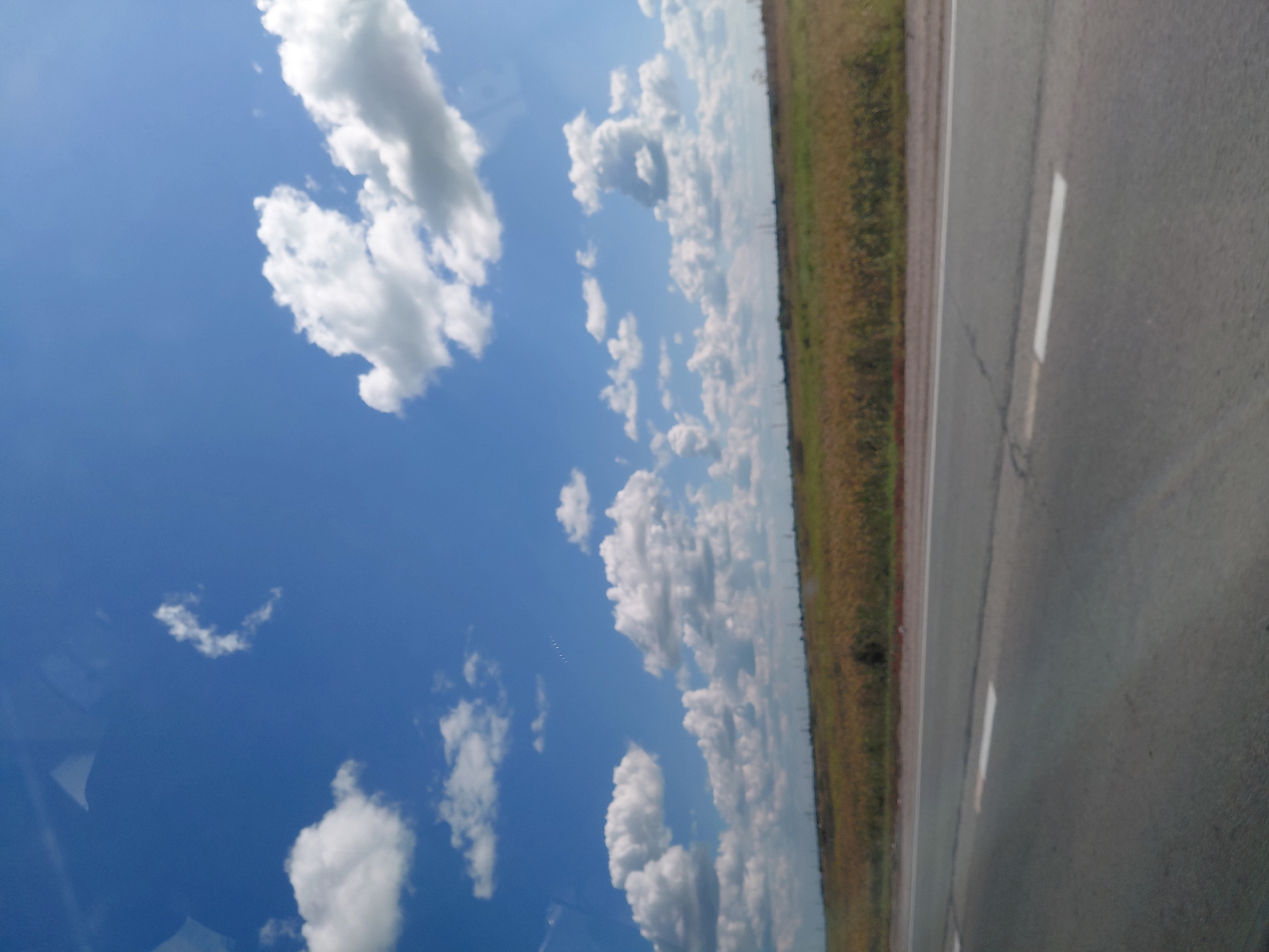 The image shows a highway with a field and a blue sky with clouds in the background. The highway is paved and has white lines marking the lanes. The field is covered with dry grass and some green plants. The sky is clear with a few white clouds. The scene is likely taken on a sunny day. There are no people or any other objects visible in the image. The location is not specified in the image itself, but the question mentions the city of Chulym, Russia.