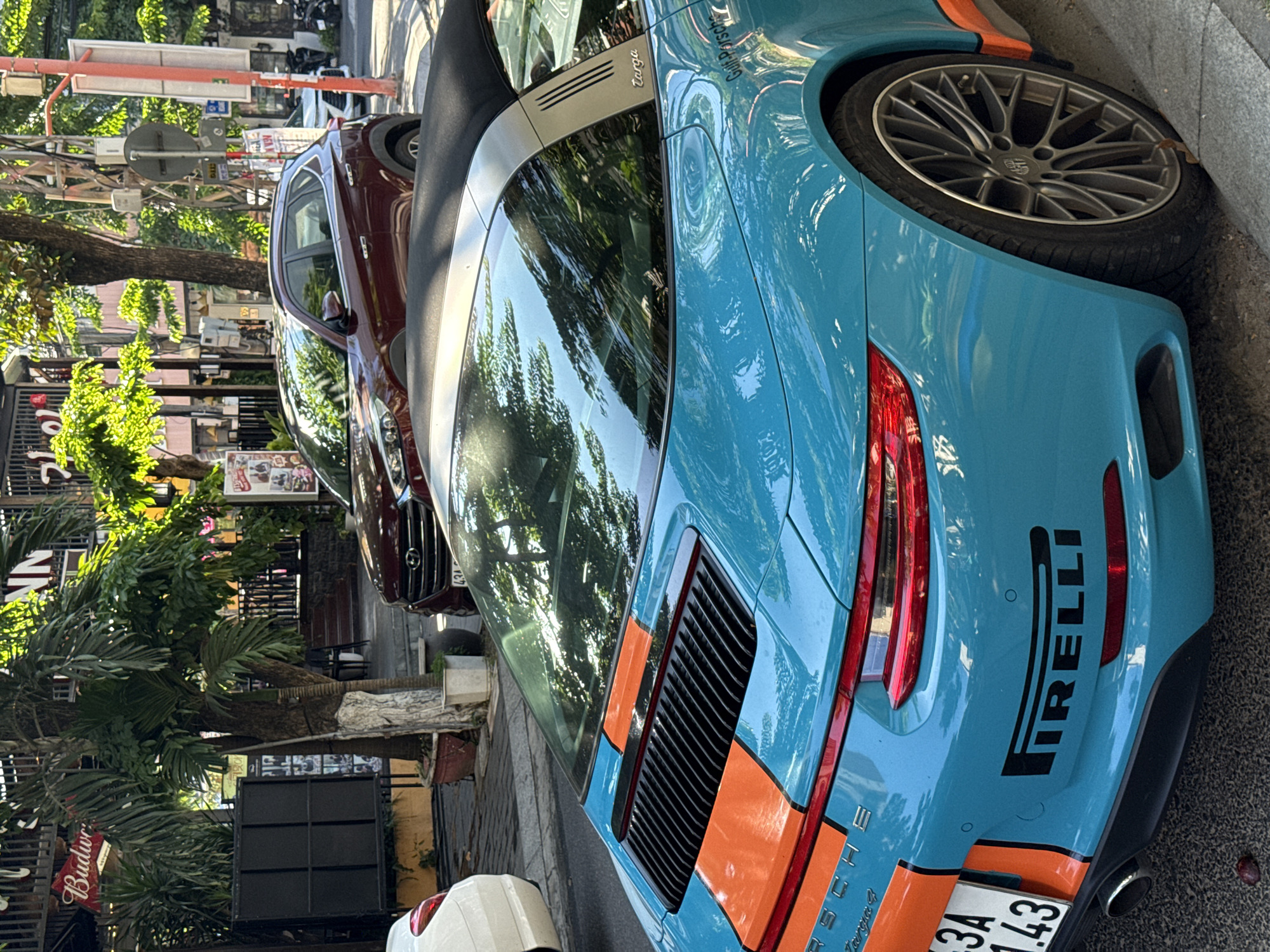 The image shows the rear of a blue and orange Porsche Targa 4 parked on a street in Đà Nẵng, Vietnam. The car features a large Pirelli logo and a license plate with the code "3A 143." The car is parked next to a curb on a street lined with trees and buildings. Reflections of trees and buildings are visible on the car's rear window. To the left is the exterior of a bar with a Budweiser sign. Behind the Porsche is a maroon-colored car. The general environment is a city street during daylight hours.