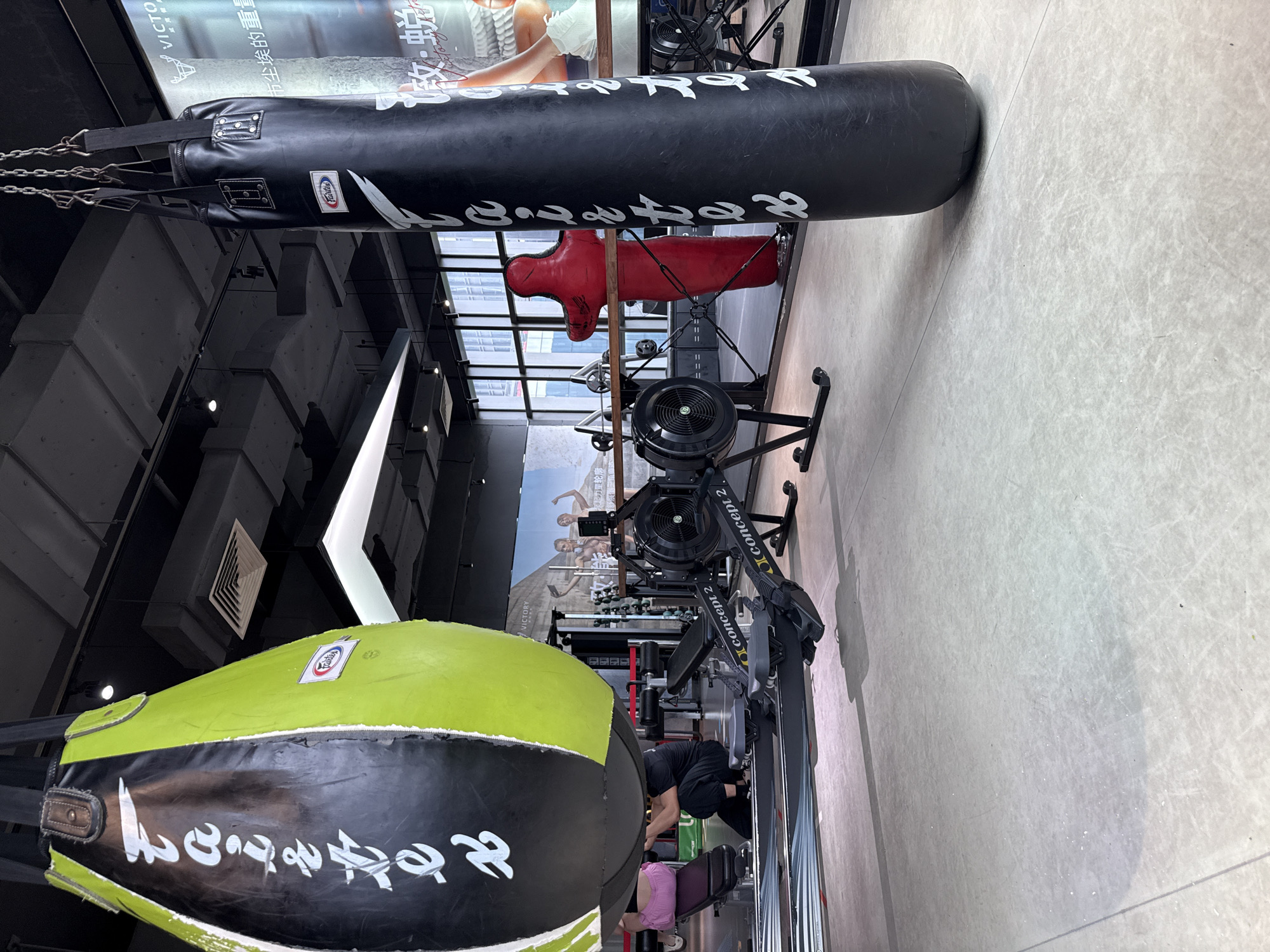 This is a medium shot of a gym in Haizhu District, China, featuring various exercise equipment. Two large punching bags hang from the ceiling; one is black, and the other is a combination of lime green and black. In the mid-ground, two rowing machines are visible, labeled "Concept 2." To the right of the rowing machines, a red, full-body dummy used for combat training stands upright. In the background, a large poster is displayed, featuring a deer logo and Chinese text. A woman in a pink top is seated on a weight bench to the left of the frame, and a person in black athletic wear is kneeling beside her. The environment appears to be a modern fitness facility with polished concrete flooring and industrial-style ceiling elements.