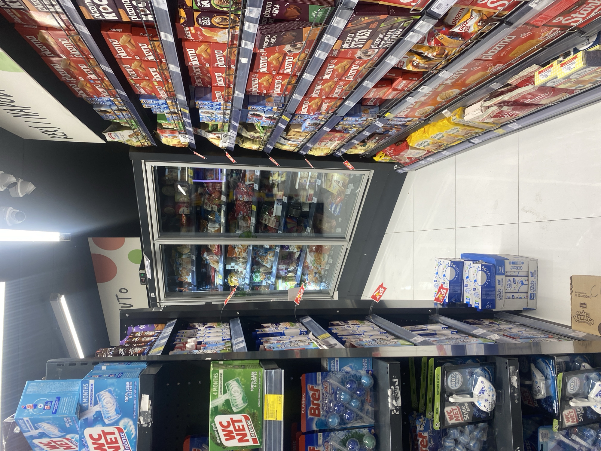 The image shows an aisle in a grocery store. On the top shelves, there are various types of “plazma” brand biscuits, including flavors like “RolS” and “STAKS”. Further down the aisle, a refrigerated section displays various frozen food products. Below the freezer, there are shelves stocked with bathroom cleaning supplies, including “WC Net” and “Bref” toilet bowl cleaners. Boxes of “Mleko” milk are also visible. Based on the writing on the products and the price tags, it appears to be located in a grocery store in Budva, Montenegro.