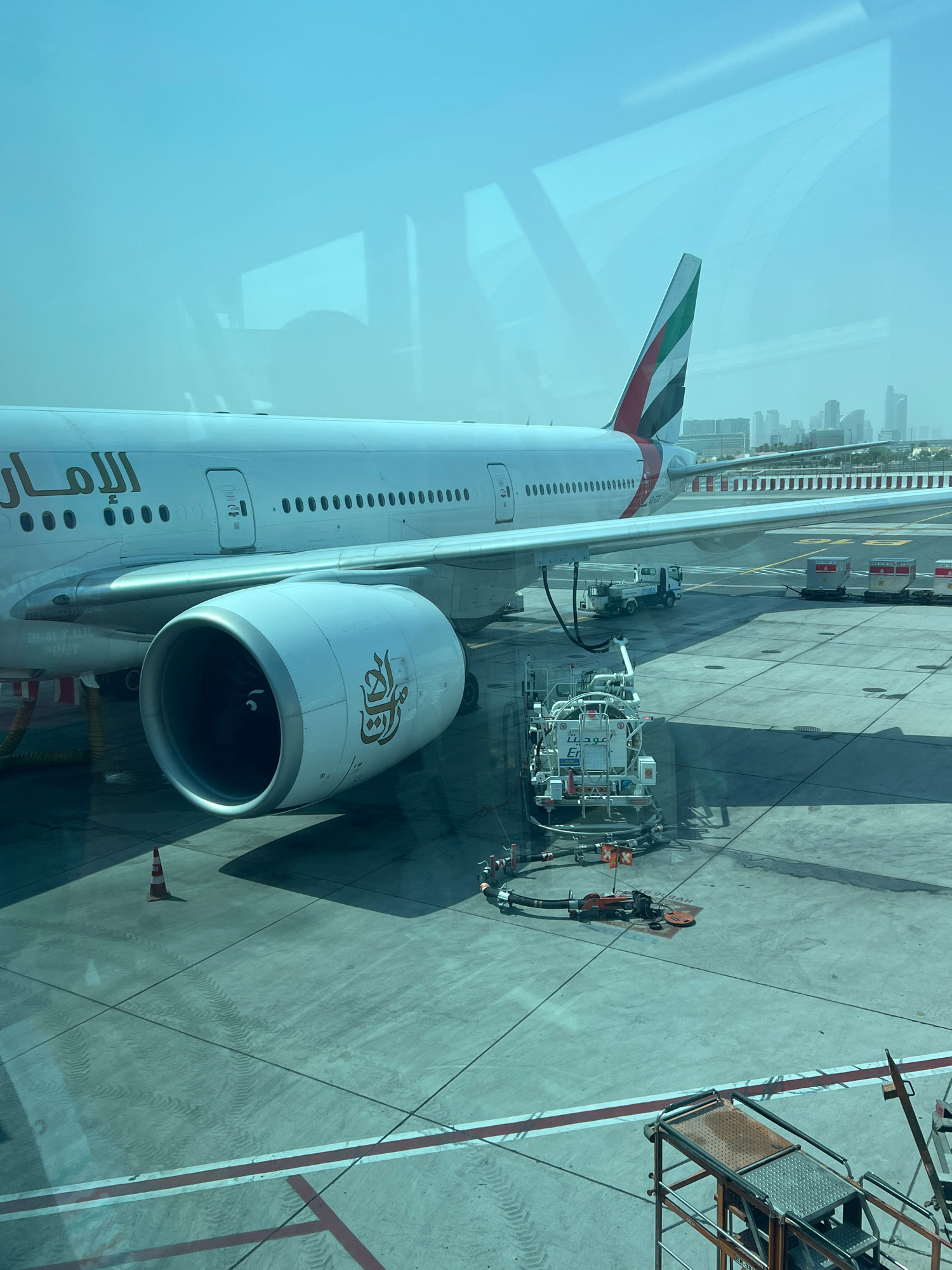 An Emirates airplane is parked at an airport gate in Dubai, United Arab Emirates, possibly Dubai International Airport (DXB). The airplane is a wide-body jet, likely a Boeing 777, indicated by its large engines and the airline's livery. The tailfin displays the UAE flag colors: red, green, white, and black. The aircraft's fuselage has "الإمارات" (Emirates) written in Arabic script.
The airplane is being refueled from a white fuel truck with Arabic text visible on its side. The fuel hoses are connected to the aircraft's underside. In the background, other airport vehicles, including baggage carts, are visible on the tarmac. The city skyline of Dubai, with its characteristic modern skyscrapers, can be seen in the distance, suggesting the airport's urban location. The scene is captured through a window, showing reflections of the sky and possibly the interior of the terminal on the glass. The overall environment is an airport tarmac on a clear, sunny day. There are no people prominently visible in the image.