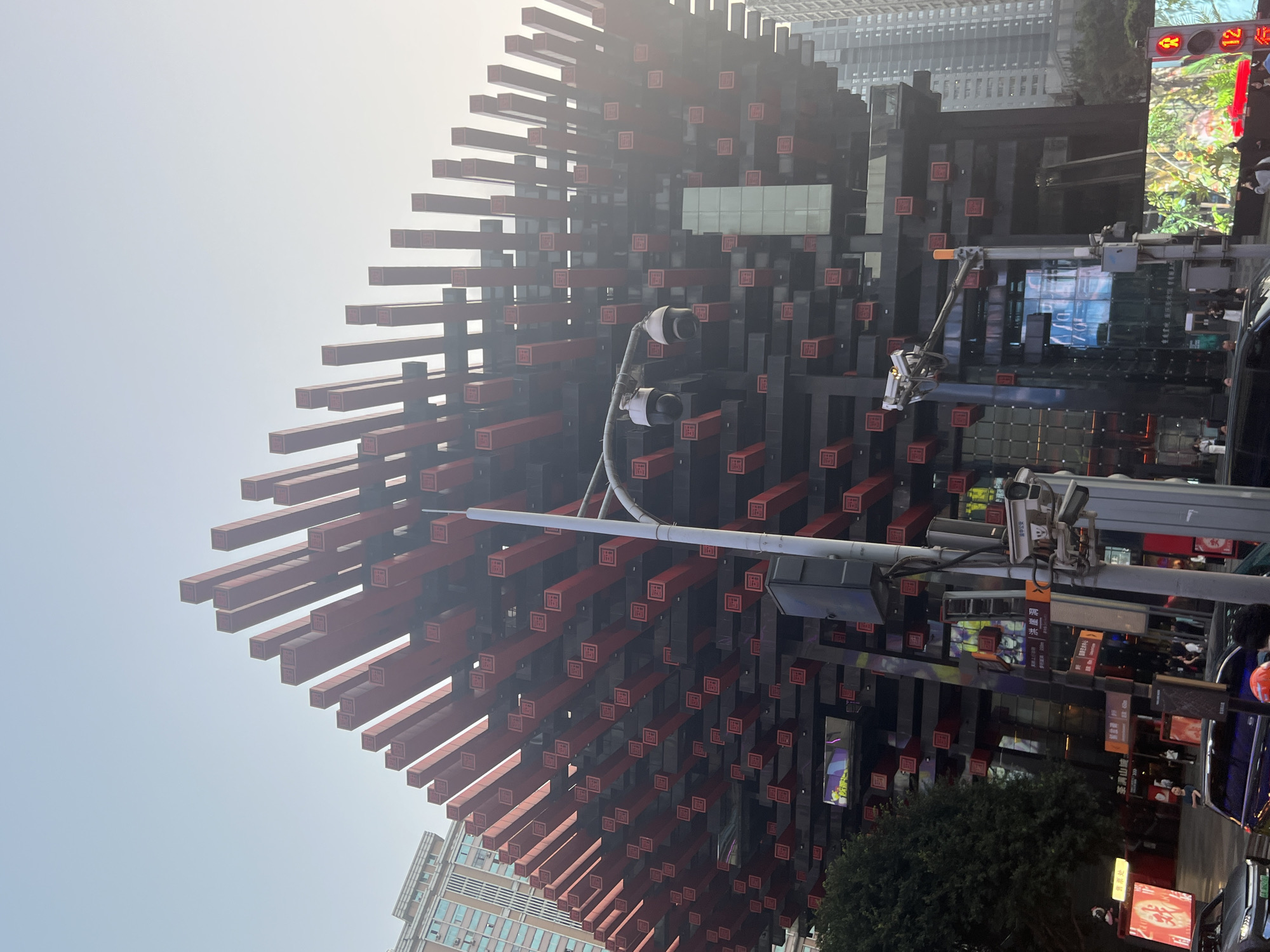 The image depicts a close-up, low-angle view of the Raffles City Chongqing complex, a landmark building located in Yuzhong District, Chongqing, China. The complex's distinctive architectural design is characterized by numerous dark grey horizontal beams protruding from the main structure. Each beam is adorned with a red square containing Chinese characters.
A surveillance camera is mounted on a pole, positioned near the center of the frame. Below the beams, there is an active street scene with various commercial signs and digital billboards, though the people and moving vehicles are blurred. A small tree is partially visible in the lower right. The sky is overcast with light.