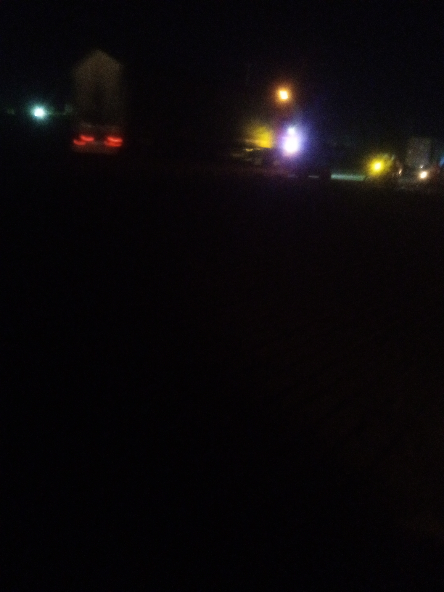 The image is a blurry, dark photograph taken at night. It appears to be an outdoor scene with several lights visible. In the upper left, there is a bright white light, and to its right, a building with red lights, possibly taillights of a vehicle. Further to the right, there are other lights, including a yellow light, a purple light, and a bright white light, suggesting streetlights or other artificial illumination. The overall impression is of a street or an open area at night, possibly with vehicles and buildings in the distance. The image lacks clear details due to the low light conditions and blurriness. No people or specific landmarks that would indicate Nassarawa, Nigeria are discernible.