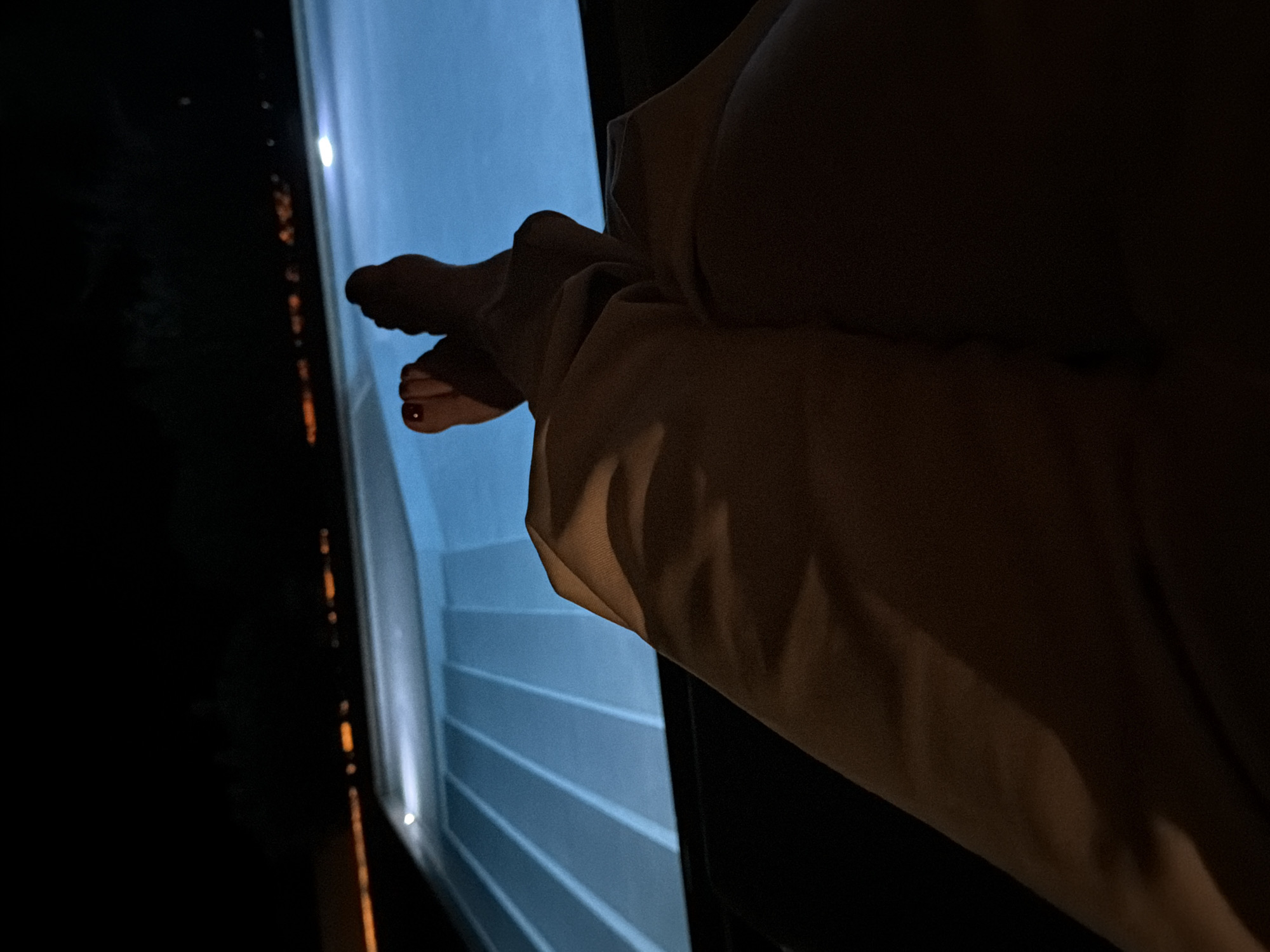 The image shows a close-up view of a person's legs and feet, illuminated by artificial light, with a swimming pool in the background. The person is wearing light-colored pants that are draped over their legs. Their feet are crossed, and the toes of one foot are visible, with red nail polish on one of the toenails. The swimming pool is lit with blue underwater lights, creating a vibrant contrast with the dark surroundings. The water appears calm, with distinct lines of light reflecting on its surface, possibly indicating steps or the pool's depth. In the distance, beyond the pool, there are blurred lights that suggest a cityscape or a densely populated area, hinting at a location within or near a populated region, rather than a secluded rural setting. The overall ambiance is one of relaxation and quiet enjoyment, possibly during nighttime. The location is identified as Viglafia, Greece, although the specific characteristics of Viglafia that are discernible from the image are limited to the presence of artificial lighting indicative of a populated area.