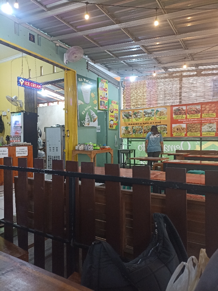 The scene is located at a restaurant at coordinates -7.425, 109.228 in the city of Purwokerto, Indonesia. The restaurant is brightly lit with yellow lights, likely fluorescent, and has an exposed ceiling with visible structural support beams. The interior walls are painted a light yellow and decorated with various posters. On the left-hand side, a refrigerator is visible, along with a sign reading "Ice Cream." On the right, a large menu board is visible. There are several tables and chairs scattered throughout the restaurant. There is a woman standing near a table, wearing a green tank top and jeans. She is facing the camera. The floor is covered in a light colored tile material and the space is enclosed with a black metal fence. Behind the fence are multiple black bags and an empty white plastic bag. The fence itself is made of vertical wooden panels. The majority of the space is taken up by tables with wooden tops and black metal legs. The tables are set with empty seats. The restaurant is empty except for the one woman standing near a table. All the furniture appears to be made of wood.