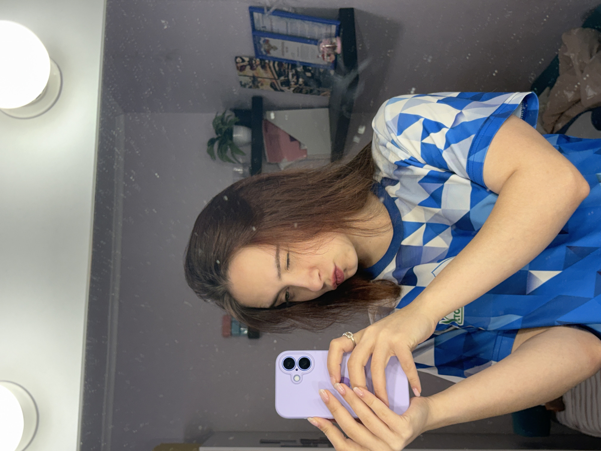 A woman is taking a selfie in front of a mirror. She is holding a purple smartphone. She is wearing a blue shirt with white geometric patterns. The mirror shows a reflection of a shelf with decorations and a plant. There is also a white vanity light fixture next to the mirror. The location is not identifiable based on the image.