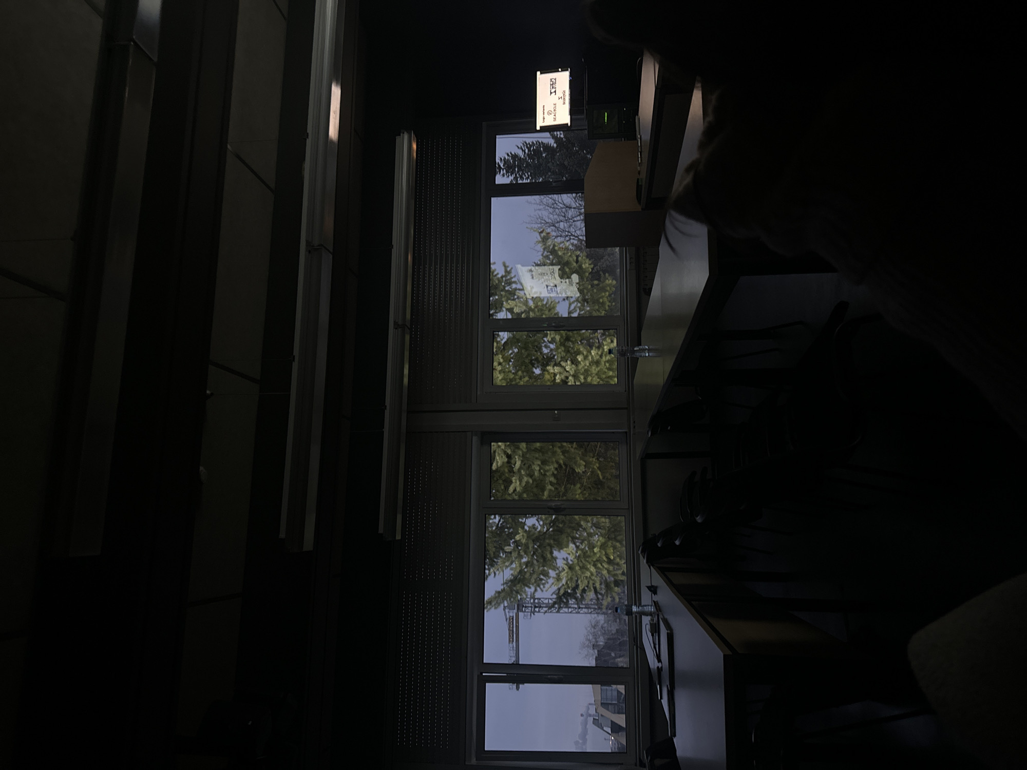 The image depicts an interior view through a multi-paned window. Outside, a building and trees are visible. A crane can be seen in the distance. Inside, part of a desk and seating are visible, along with what appears to be a sign or screen. The location is likely Sofia, Bulgaria, based on the signage displayed.