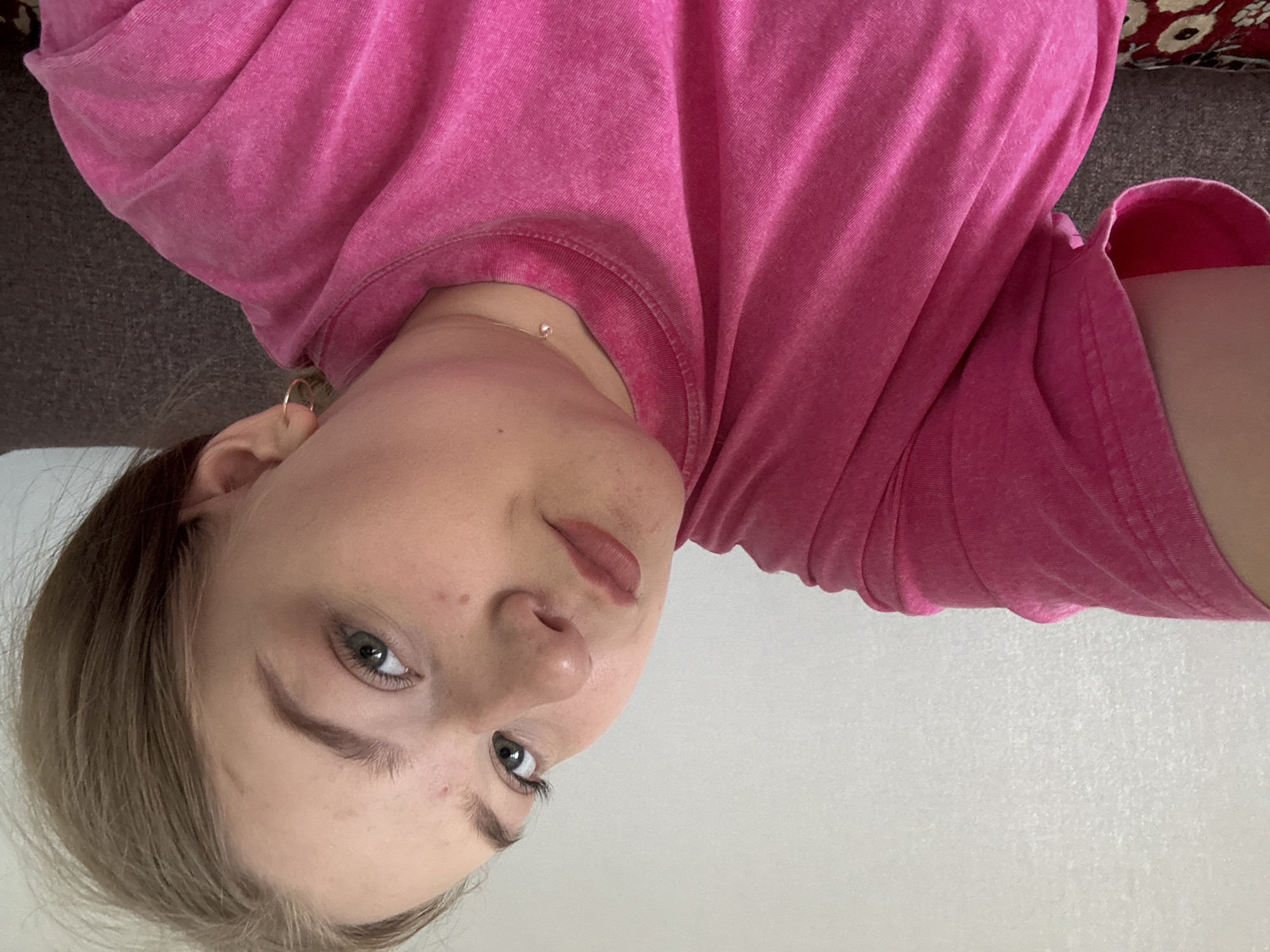 A close-up selfie shows a young woman with fair skin, blonde hair, and blue eyes. She is wearing a pink t-shirt and a delicate gold necklace. Her lips are a natural pink shade. She appears to be indoors against a backdrop of a textured white wall. Part of a grey couch and a patterned red cushion are visible in the upper left corner of the frame. There is no information indicating that the image was taken in the city of Фоки, Russia.