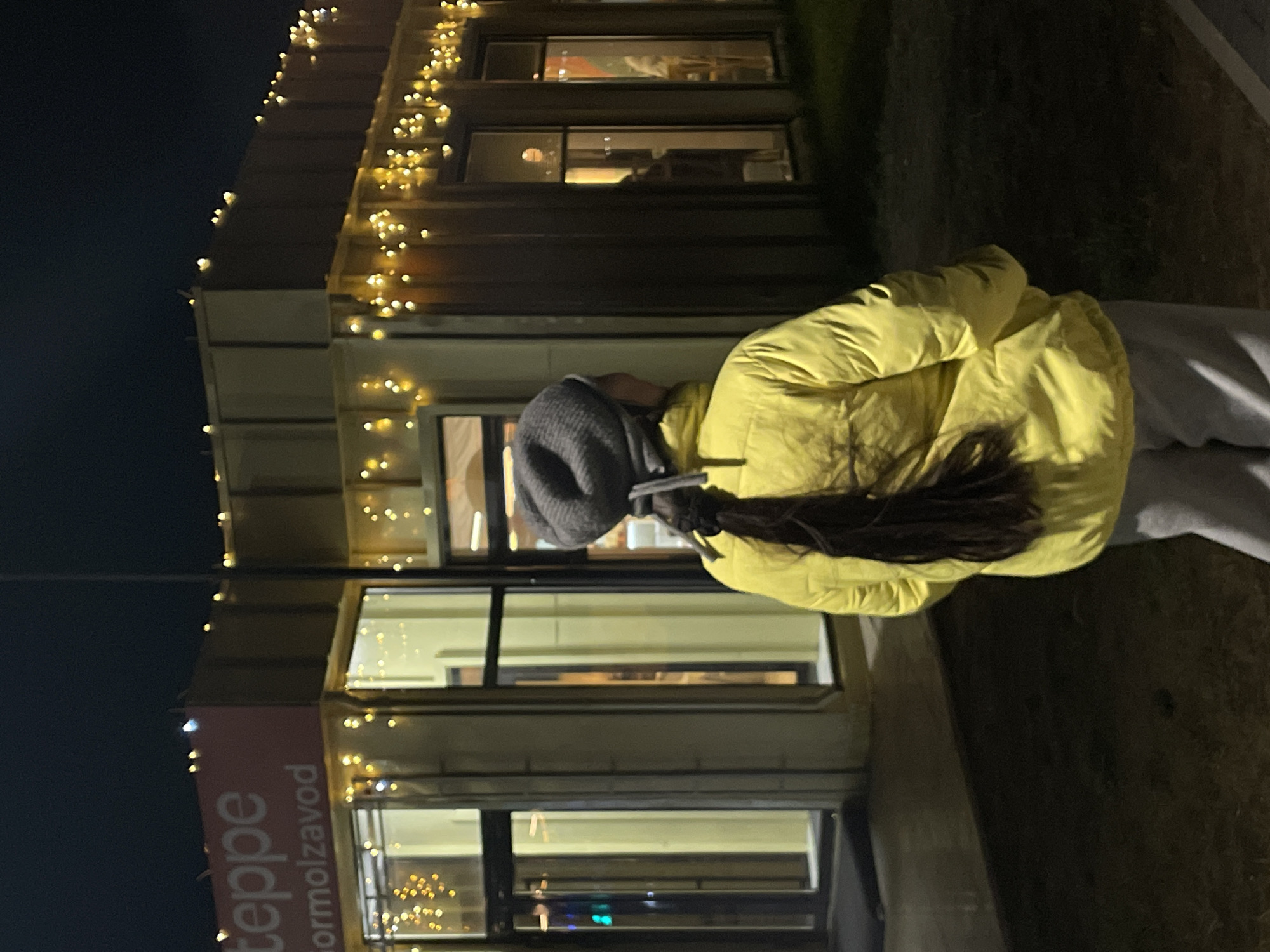 A building in Kokshetau, Kazakhstan, is decorated with strings of lights. The building has a sign in Cyrillic script reading "teppe Formolzavod." A person wearing a yellow jacket, grey pants, and a grey hat stands in front of the building. The photo was taken at night.