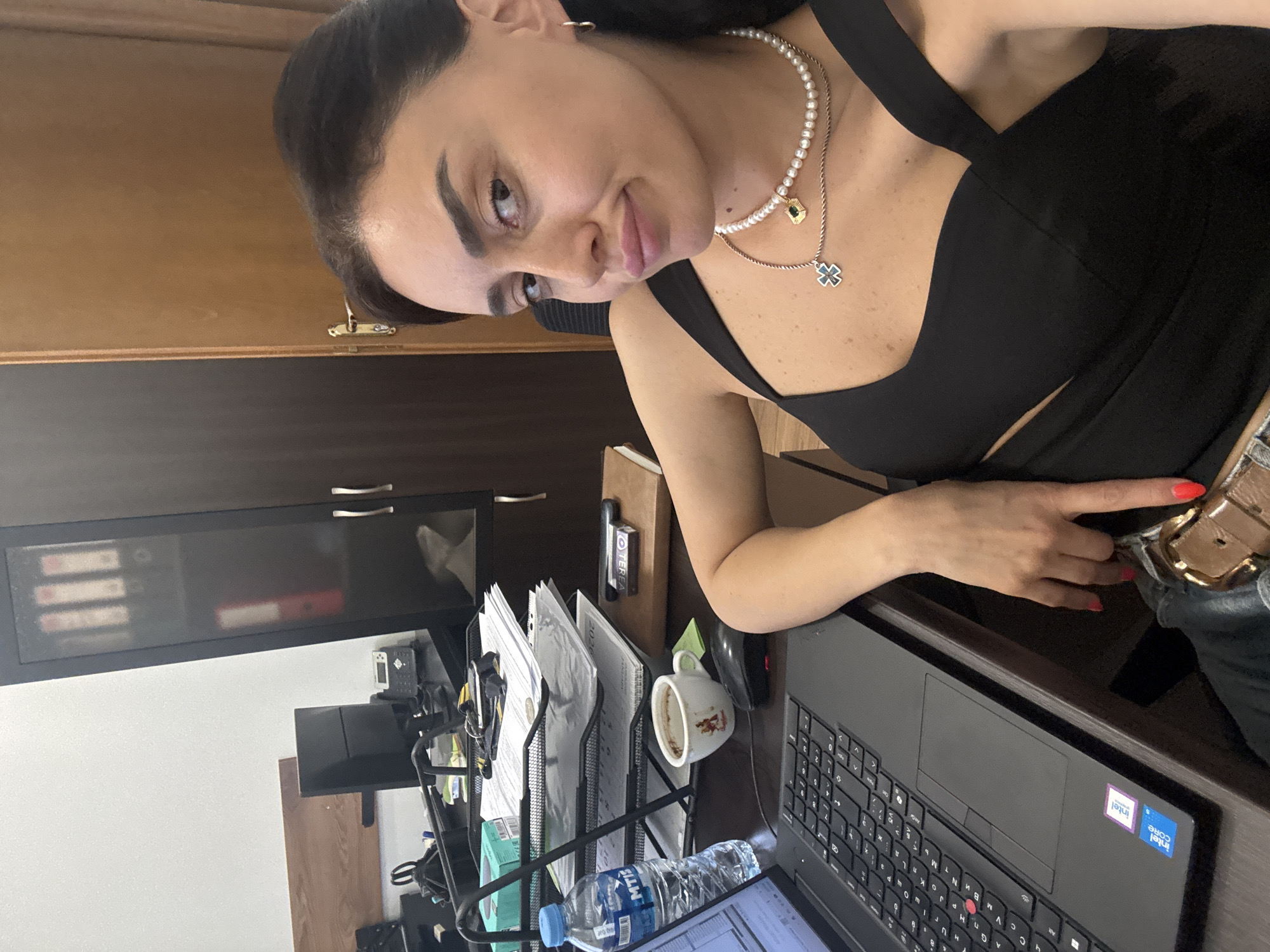A woman with dark hair and dark eyes is taking a selfie in an office. She is wearing a black top with a low neckline and a pearl necklace with two pendants, one of which is a green gemstone. She has on hoop earrings and bright red nail polish. Her left arm is resting on the desk, and her right hand is visible near her lap, also with red nail polish.
The desk is cluttered with office supplies, including a laptop, a bottle of water, a coffee cup, a mouse, and a tray filled with papers. The background shows a dark wood cabinet with binders on the shelves, a wooden door, and a white wall. The overall setting appears to be an office environment. The presence of a laptop and office supplies suggests a workspace. The woman's attire, a selfie in an office setting, and the presence of a laptop suggest she may be working or taking a break from work. The image does not contain any specific visual cues that can be used to determine the city or country.
