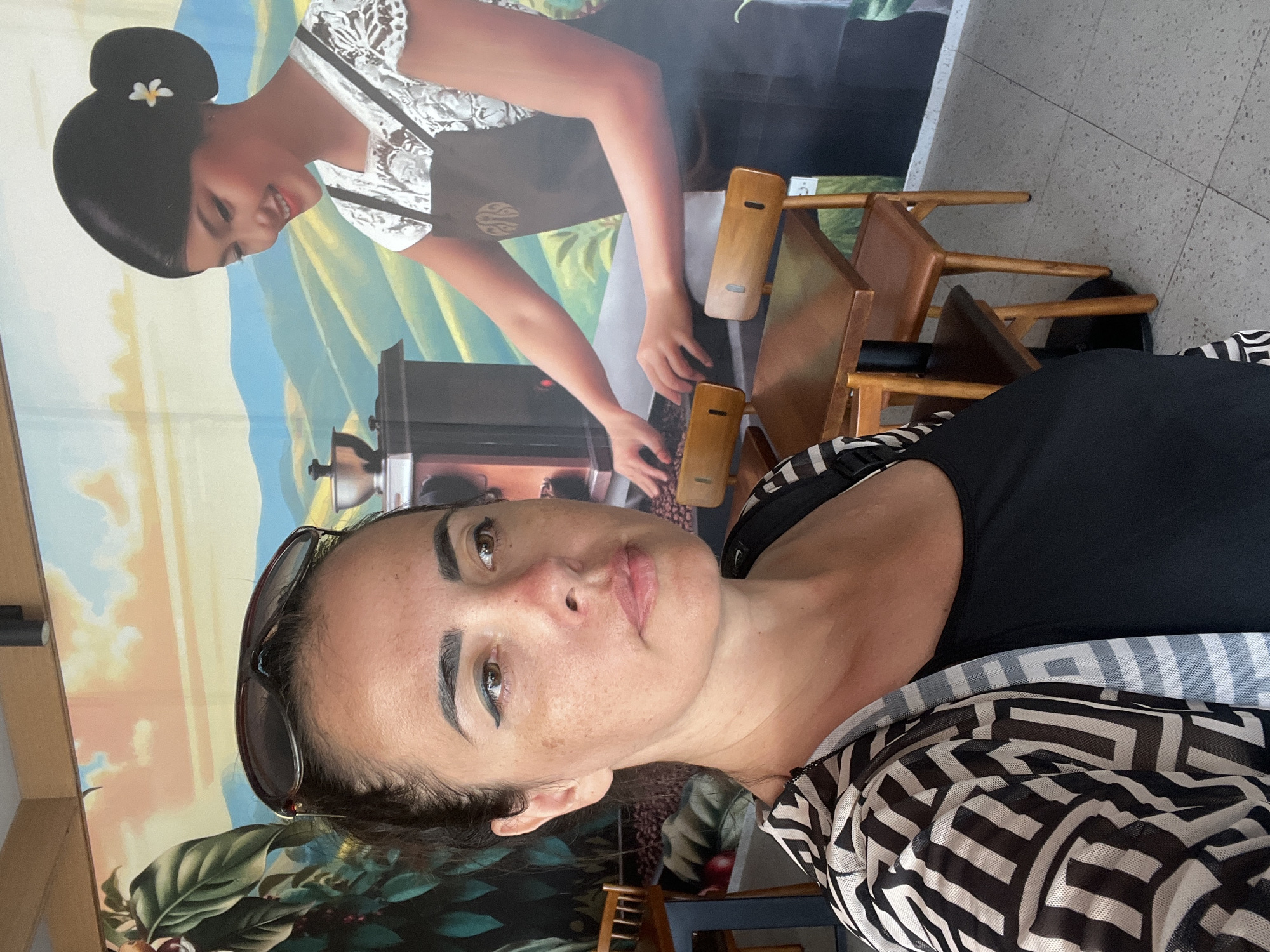 In Kuta Selatan, Indonesia, a woman poses for a selfie in front of a vibrant mural depicting a coffee plantation and a woman preparing coffee. The woman in the foreground has dark hair, brown eyes, and is wearing sunglasses perched on her head. She has a patterned kimono-style cover-up over a black top. The mural behind her shows lush green hills, a coffee grinder, and a smiling woman with her hair in a bun adorned with a white flower. Wooden tables and chairs are visible in the foreground and mid-ground, suggesting a cafe setting. The floor is tiled with a speckled pattern.