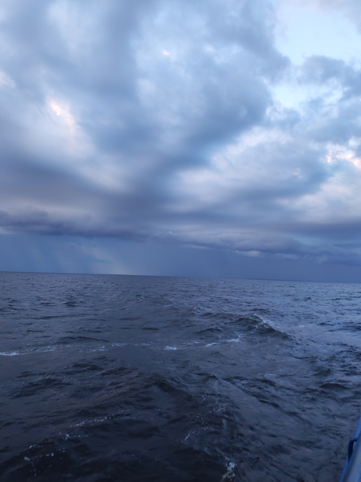 An overcast sky looms above a dark, choppy body of water, likely a sea or large lake. The clouds are heavy and gray, with streaks of lighter sky visible through breaks, suggesting either the early morning or late evening. In the distance, a subtle curtain of rain or mist falls from the clouds to the water. The water itself is a deep blue-gray, with whitecaps and ripples indicating a moderate wind. The scene is devoid of any visible human presence or structures, conveying a sense of vastness and natural power. The location is Unknown, Russia.