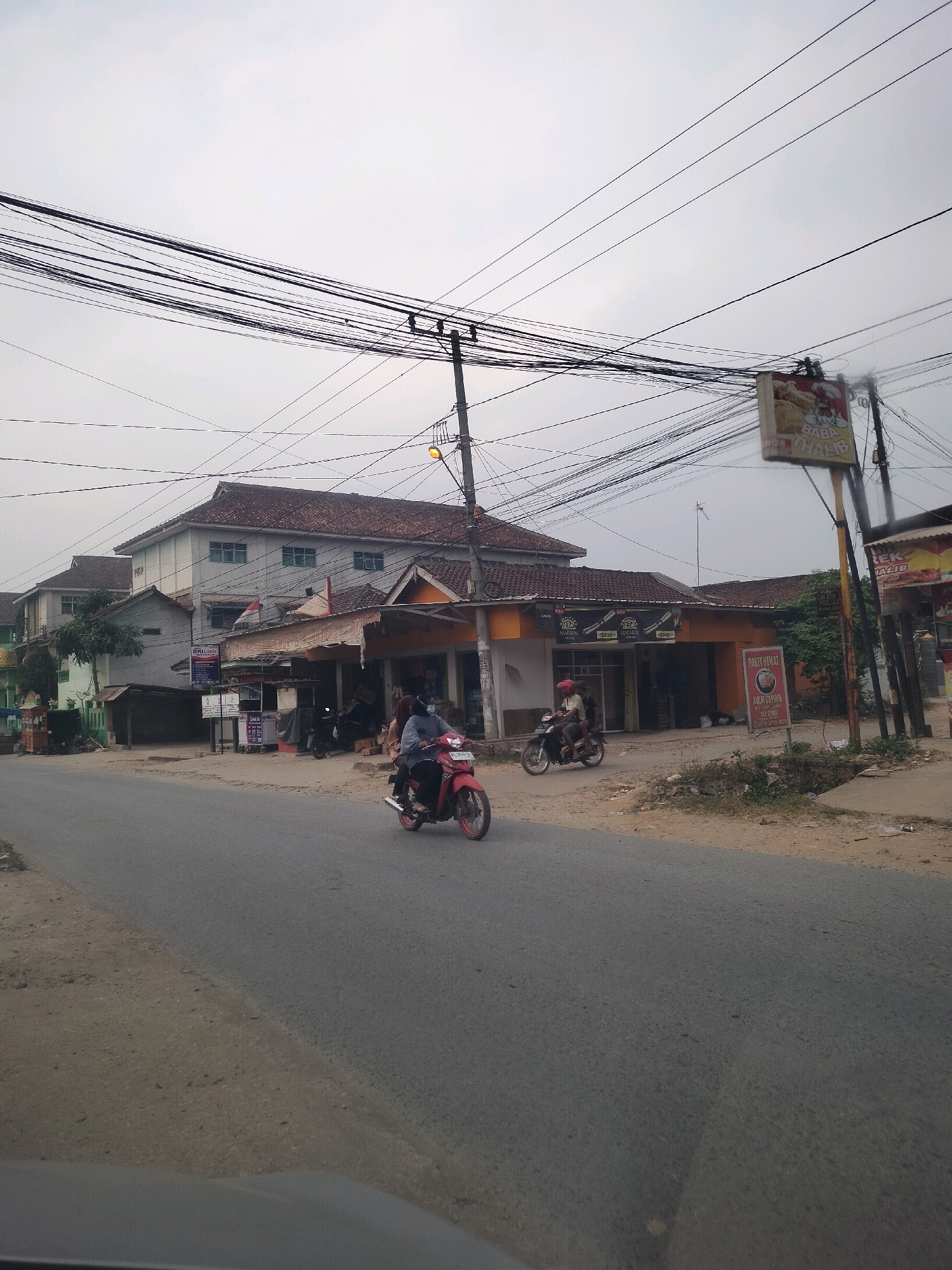The image shows a street scene in Wayakroy, Indonesia, with a road in the foreground and buildings lining the street on the right. A motorcycle with two people is traveling on the road. The buildings appear to be a mix of commercial establishments and residential units, with awnings and signage visible. Overhead, a dense network of power lines stretches across the sky, supported by utility poles. The sky is overcast, indicating a cloudy day. The general atmosphere is that of a typical urban street in Indonesia.
