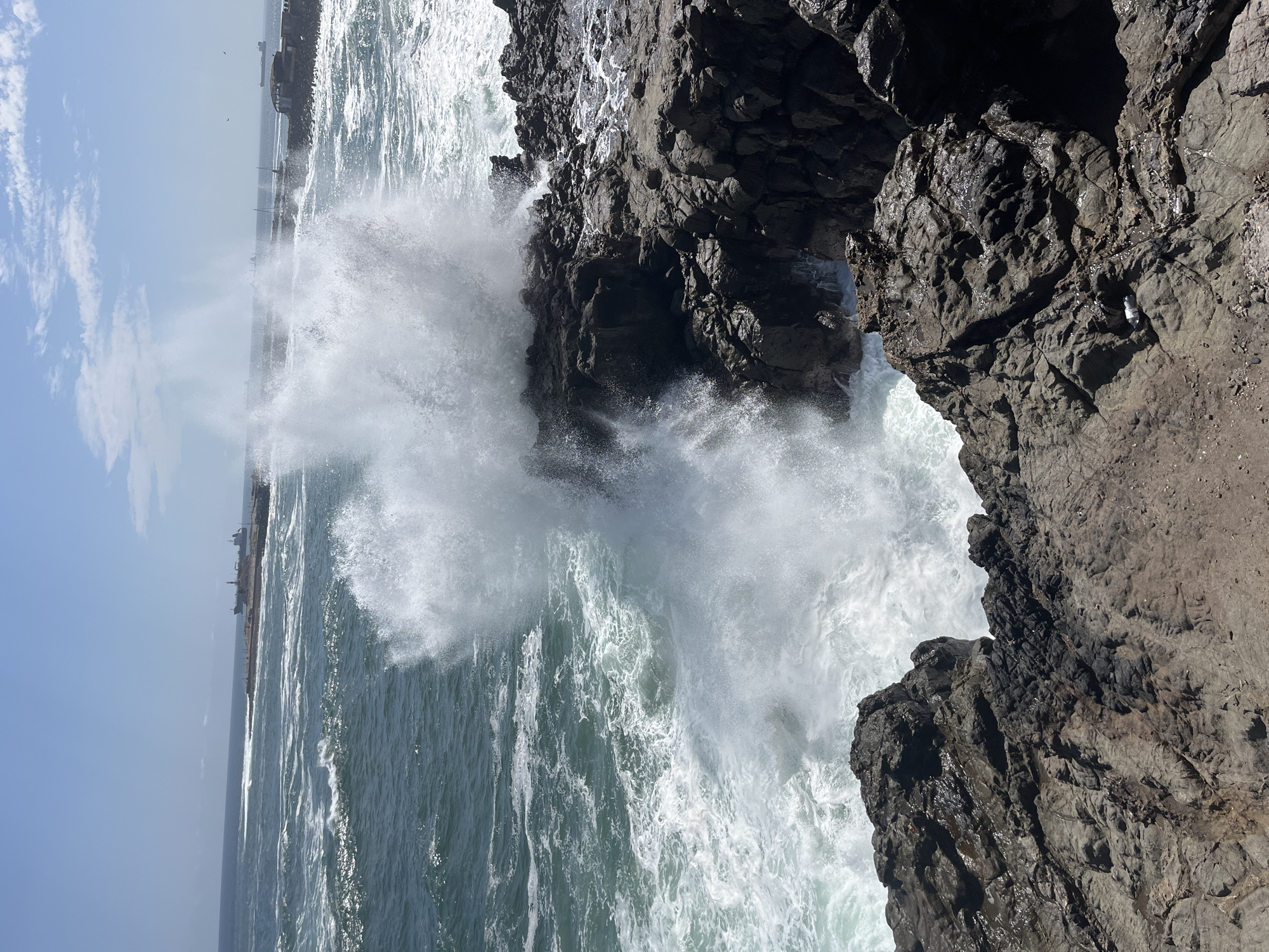 The image depicts waves crashing against the rocky coast of Arica, Chile. The view is from an elevated position, possibly on the cliffs, looking out over the ocean. Large waves are breaking with considerable force, sending spray high into the air, which obscures some features of the coastline. In the distance, a low-lying landform is visible, with some indistinct structures that may be docks or buildings, partially obscured by the spray from the waves. The cliffs in the foreground are dark and rugged. The sky is mostly clear blue. The photograph appears to have been taken during daylight.