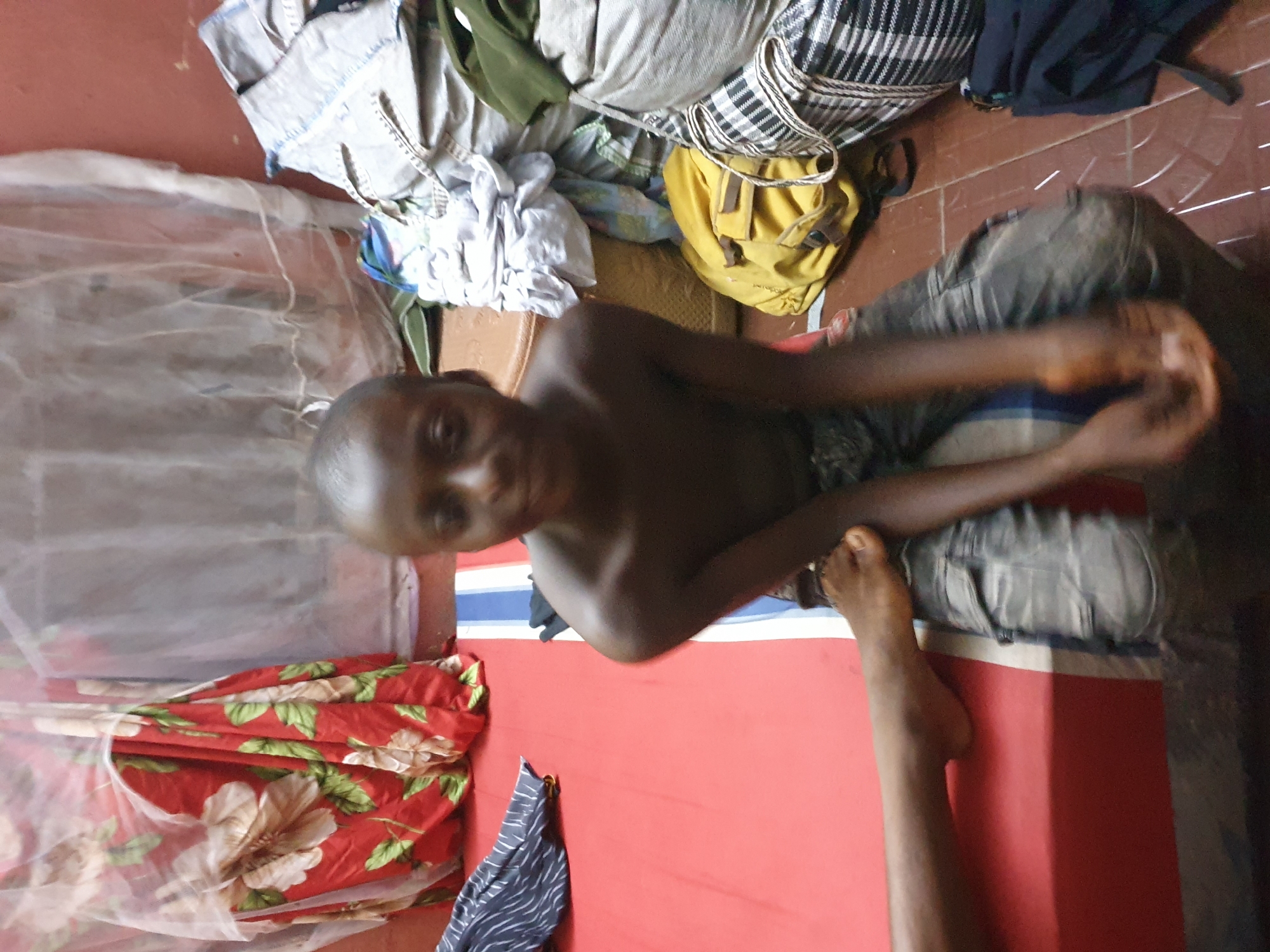The image captures a young, dark-skinned person, likely a boy, sitting indoors and looking directly at the camera with a neutral expression. He is shirtless and wears dark trousers, seated on a red mattress or mat placed on a reddish-brown tiled floor. His hands are clasped in front of him.
To the boy's left, a white mosquito net is draped over what appears to be a sleeping area. A red fabric with large white and green floral patterns is neatly folded on the mattress, below the mosquito net. Another piece of fabric, dark blue with white stripes, lies on the mattress in the lower left foreground.
Behind the boy, various clothes and personal items are piled up on the floor or an unseen surface. These include light-colored garments, a green fabric, a black and white striped bag or textile, and other dark blue clothing. A yellow backpack with brown straps is also visible among these belongings. Part of a bare foot, possibly belonging to another person, is visible on the right side of the frame, resting near the edge of the mattress.
The setting suggests a simple indoor living space or bedroom in Idah, Nigeria. The lighting is moderate, indicating either daytime or an illuminated evening. No visible text is present in the image.