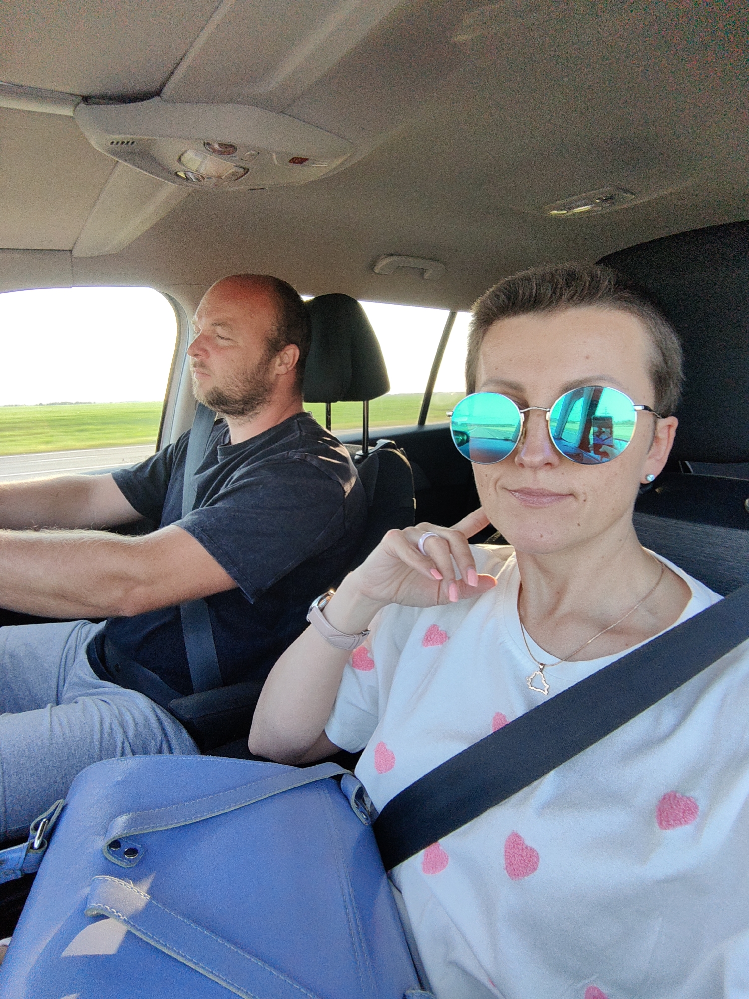 Two individuals, a man and a woman, are seated inside a car. The man, positioned in the driver's seat, is wearing a dark t-shirt and grey pants, and he has his eyes closed. The woman, in the passenger seat, is wearing a white t-shirt with pink heart patterns, and she is looking at the camera, wearing large, round, mirrored sunglasses. A blue bag is visible in the foreground. The environment outside the car appears to be a flat, open landscape with green fields, suggesting a rural or suburban setting. The presence of a seatbelt on both individuals indicates they are traveling by car. The lighting suggests it is daytime, possibly late afternoon due to the warm tones. The image appears to be taken from the perspective of someone in the back seat, looking forward at the occupants.