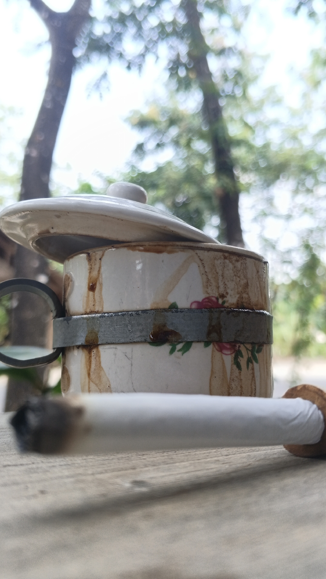 A lit cigarette rests on a wooden surface in front of a ceramic container with a lid. The container is cylindrical and appears to be old, with brown stains and a floral pattern. A metal band encircles the middle of the container. In the background, out of focus, are trees and a bright sky, suggesting an outdoor setting. The location is identified as Ngampel, Indonesia. There are no people visible in the image.