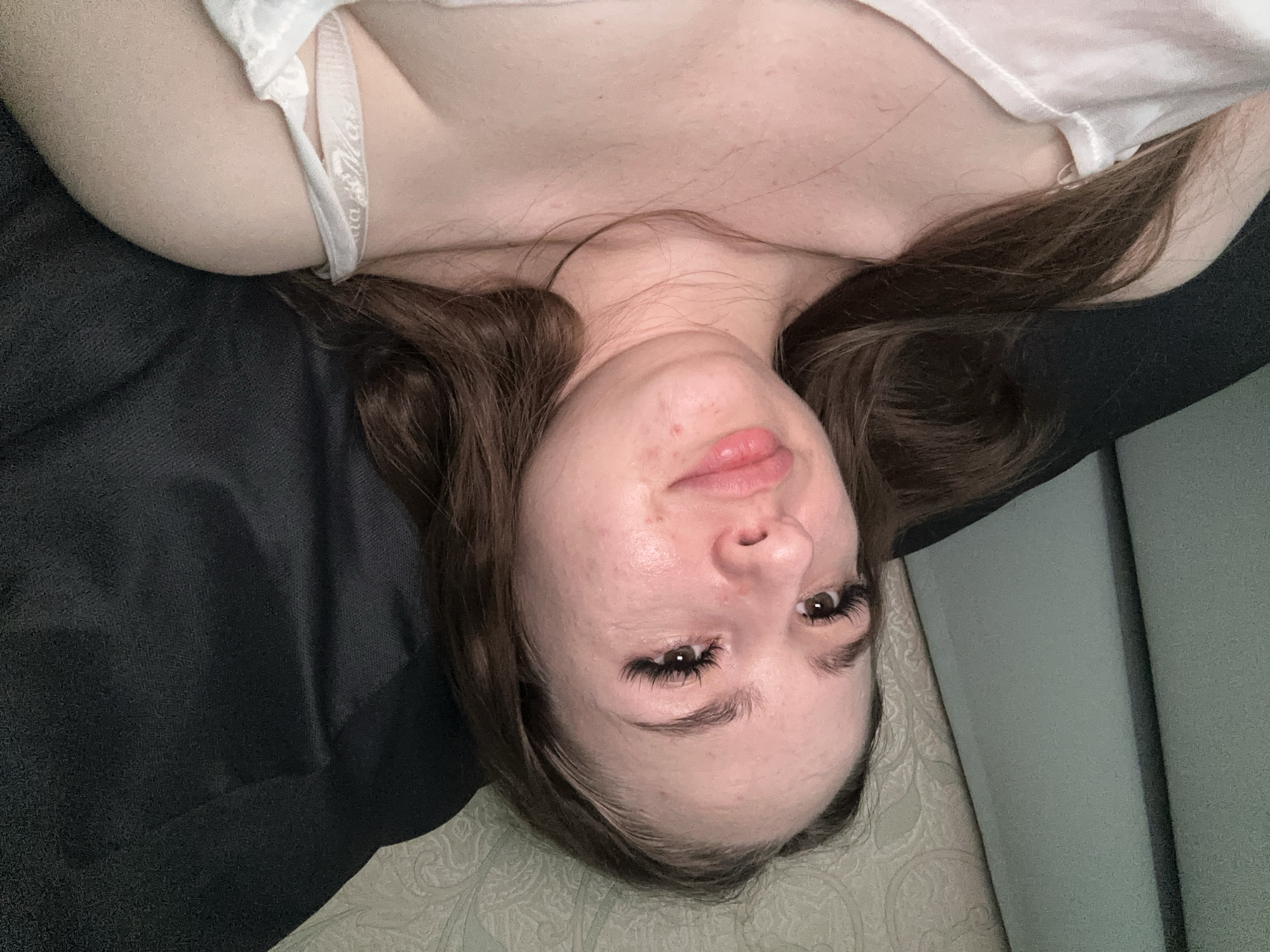 The media file is an upside-down, close-up, eye-level shot of a young person lying on their back. The person has long, dark brown hair that falls around their face and shoulders. They are wearing a white tank top with a white bra visible underneath. The background consists of dark fabric and a light-colored, textured surface, possibly a bed or couch.
The person's face is the primary focus, with their eyes open and looking upwards. They have long, dark eyelashes and their eyebrows are well-defined. Their lips are slightly parted, and there are a few small blemishes on their cheeks. The lighting appears to be indoor, with soft illumination.
There is no discernible activity or event taking place other than the person posing for the photo. The time of day and weather are not indicated. The location is unknown. The overall emotion conveyed is neutral. No visible text is present in the image.