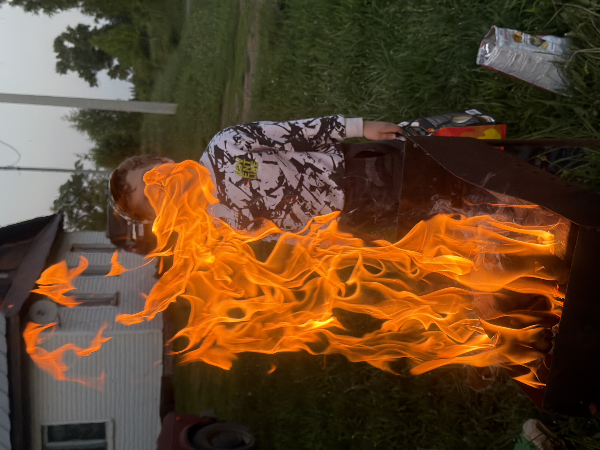 A young person in a patterned white and black shirt is near a fire in a metal fire pit. The flames are large and orange, obscuring much of what is behind them. A white house is visible to the left, partially obscured by the fire. Green grass and a few trees are present in the background. There are snack bags on the ground next to the fire pit. The picture is taken in Dubrova, Russia.