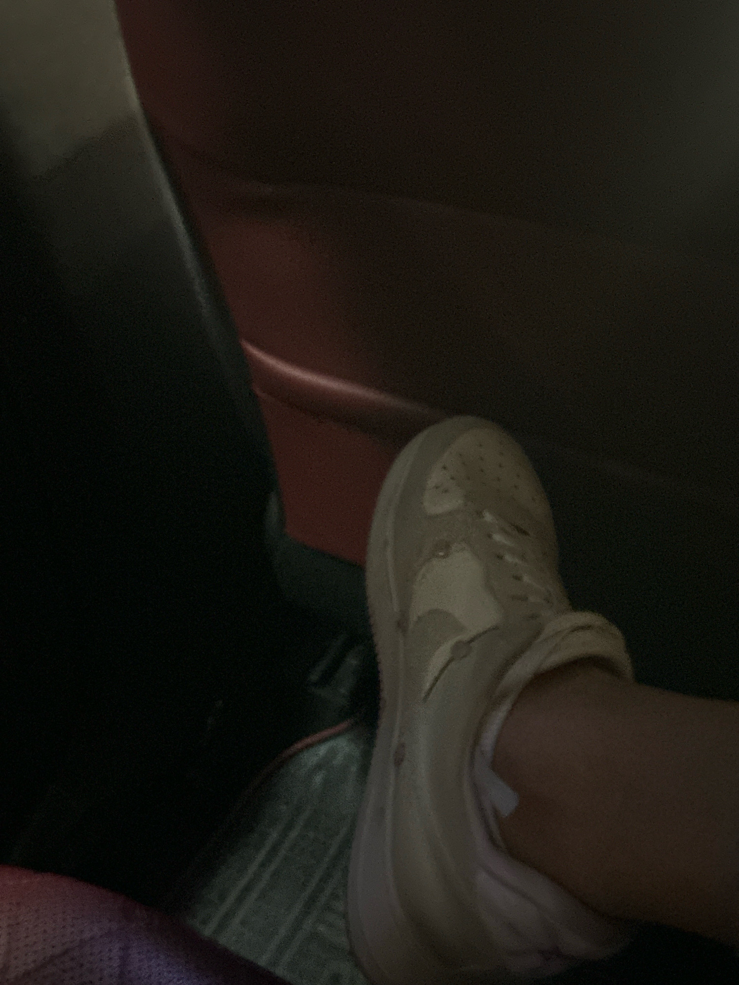 The image shows a close-up of a person's foot wearing a light-colored sneaker, likely taken inside a vehicle. The background is dark and out of focus, with hints of red and black upholstery, suggesting the interior of a car. There is no discernible location in Baku, Azerbaijan, or any other specific city mentioned in the image. The scene is static and focuses on the footwear.