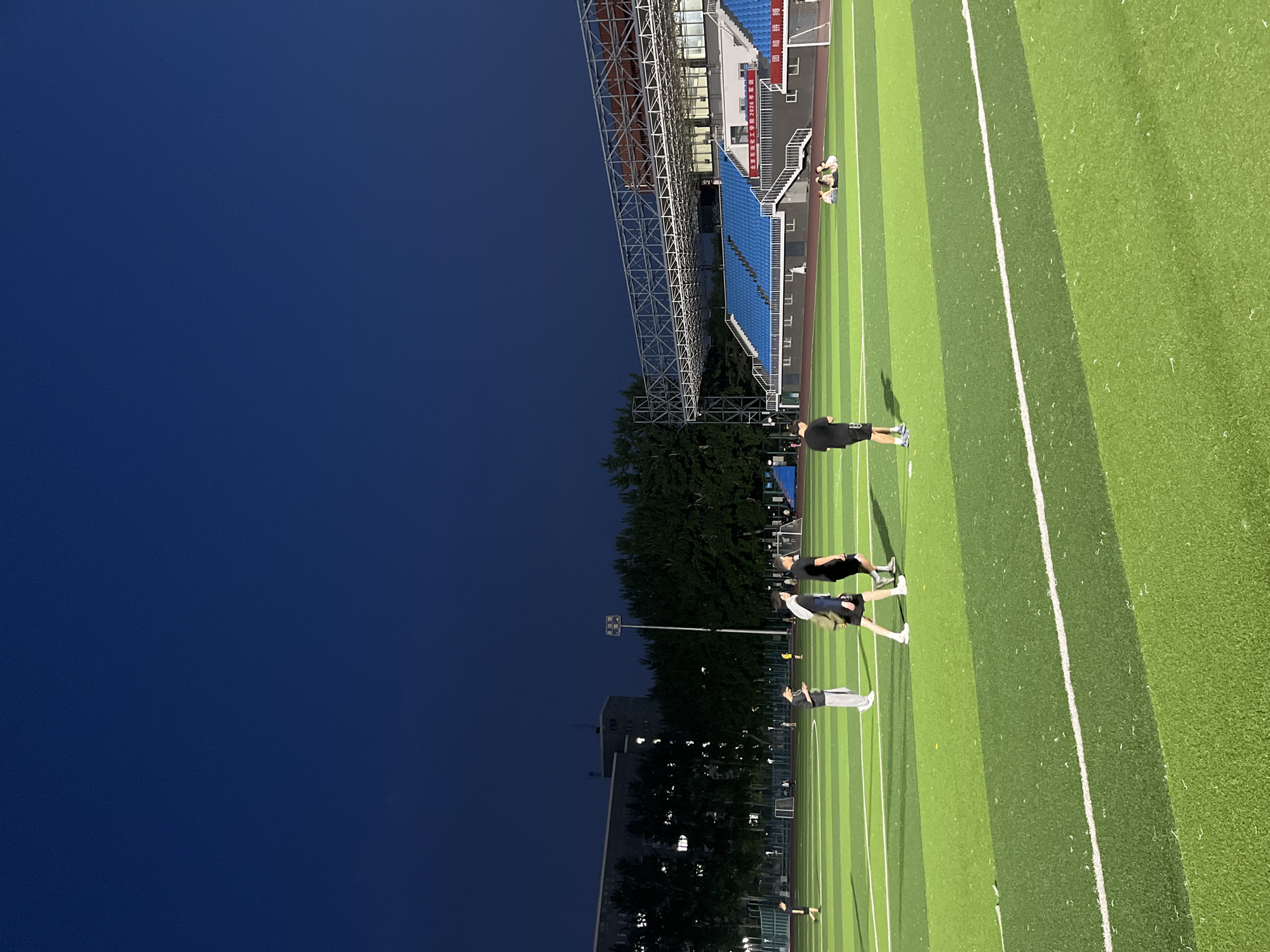 The image depicts an outdoor sports field, likely a soccer or track and field stadium, at dusk. The sky is a deep, dark blue, indicating the transition from day to night. The field itself is artificial turf, divided into alternating shades of green with white lines marking boundaries and lanes. In the background, blue stadium seating is visible, along with a metal support structure for lights or a roof. Trees line the edge of the field, and a building with illuminated windows can be seen further in the distance, suggesting an urban or suburban setting.
Several people are present on the field, seemingly engaged in physical activity. One person in a black t-shirt and shorts stands on the field, casting a shadow. Further away, a group of individuals are seen in various poses, some appearing to be stretching or resting. The overall atmosphere suggests a training session or a casual gathering after sporting events. The lighting is a mix of natural twilight and artificial stadium lights which are not clearly visible but illuminate the field.
A visible sign on the stadium seating displays Chinese characters, which translate to "Daxing District 2023 Sports Meeting" and "Tianjin University of Science and Technology". The presence of this text clearly indicates the location and the context of the event as a sports meeting.