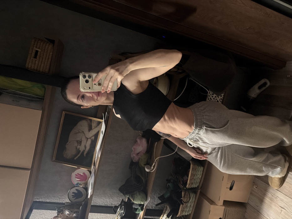 A slender woman in a black crop top and gray sweatpants takes a mirror selfie with an iPhone. Her physique is visible, highlighting her core. The background includes a woven basket, framed artwork, shelves cluttered with decorative items, and cardboard boxes. The overall scene suggests a domestic interior, possibly a bedroom or living area, with personal belongings and decor.