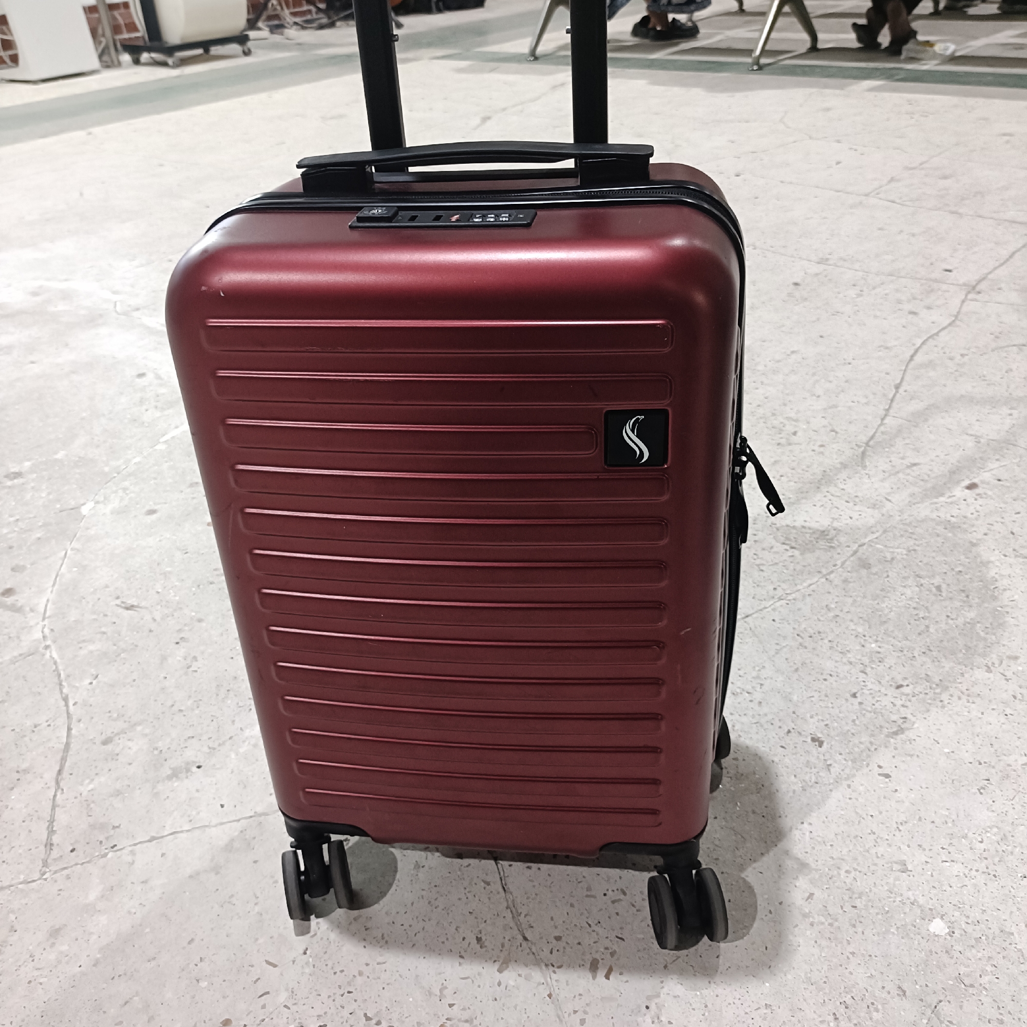 A dark red, hard-shell carry-on suitcase stands on a light-colored, cracked concrete floor. The suitcase has horizontal ridges and a black TSA-approved combination lock. A black logo, which appears to be a stylized "S", is affixed to the front of the suitcase. The suitcase has a retractable black handle and four black spinner wheels. In the background, several people are visible, some seated on chairs, and what appears to be a luggage cart. The scene suggests a waiting area, possibly an airport or train station. The lighting is neutral, with no distinct indication of time of day or weather. The overall mood is neutral, focused on the travel object. No visible text is present on the suitcase or in the immediate surroundings.