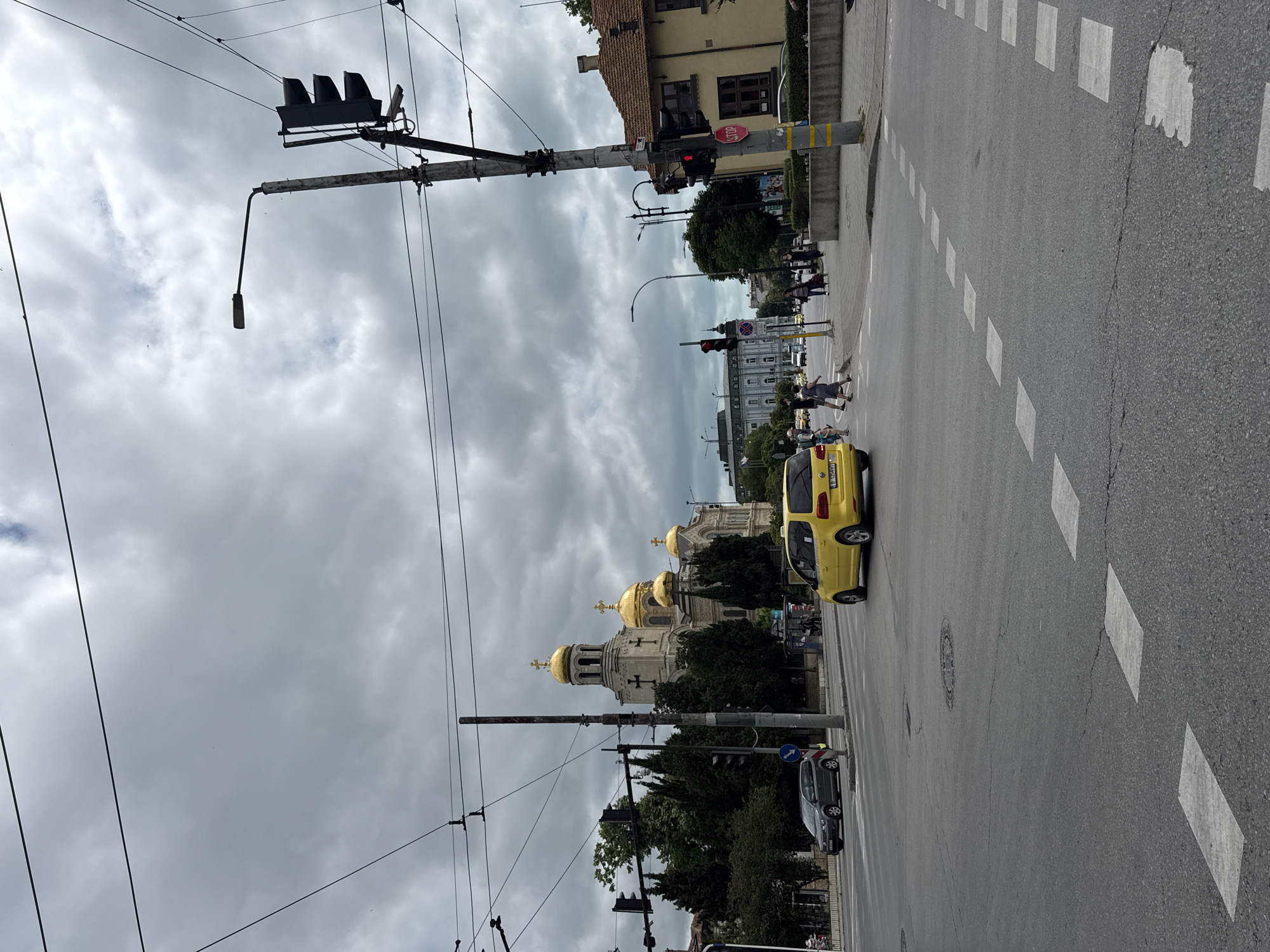 An eye-level medium outdoor shot captures a street intersection on an overcast day. In the foreground, asphalt pavement features a crosswalk with white rectangular markings and a manhole cover. To the left, a gray car drives on the road, and a traffic light is visible. A yellow taxi is positioned in the middle of the intersection, preparing to turn. Pedestrians cross the street in the mid-ground. Beyond the intersection, a large church with golden domes stands prominently. Several trees and buildings line the street, and numerous power lines crisscross the sky. A traffic light with a red illuminated signal and a "STOP" sign is attached to a utility pole on the right side of the frame.