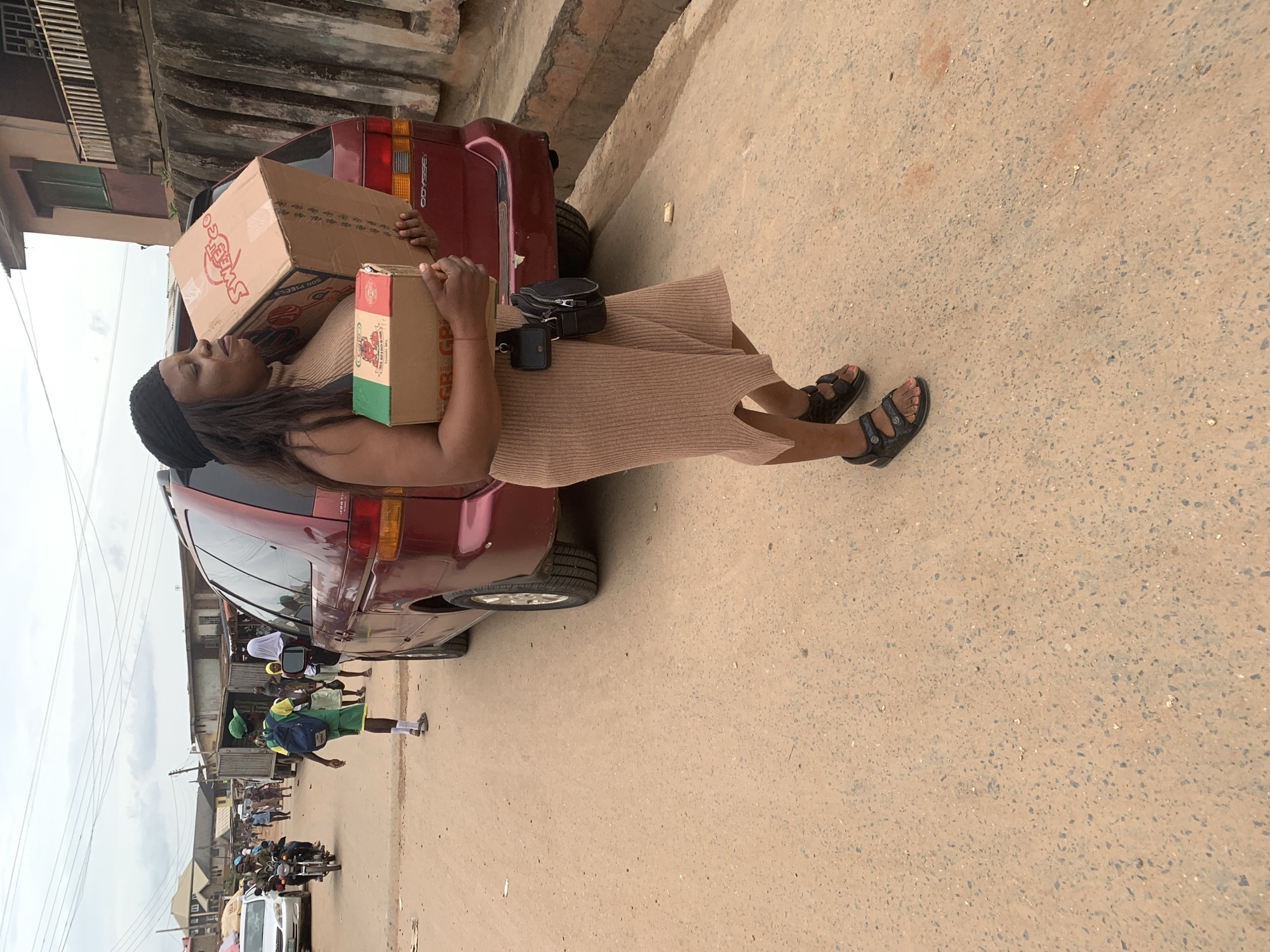 A woman is standing next to a maroon minivan, holding two cardboard boxes. She is wearing a beige dress, black sandals, and a black headscarf. The larger box is on her left, and the smaller box is in her right hand, resting on the larger one. The minivan has the word "ODYSSEY" on the rear. In the background, there are buildings and other vehicles, including motorcycles and a white car. Several people are visible, some appearing to be schoolchildren in uniform. The scene appears to be a street in Shagamu, Nigeria. The ground is paved with asphalt. The lighting suggests it is daytime, possibly overcast. The woman's facial expression is neutral, but she appears to be exerting effort to hold the boxes. Visible text on the larger box includes "Sweet Go" and "500 PIECES". The smaller box has text that includes "GR" and "GP".