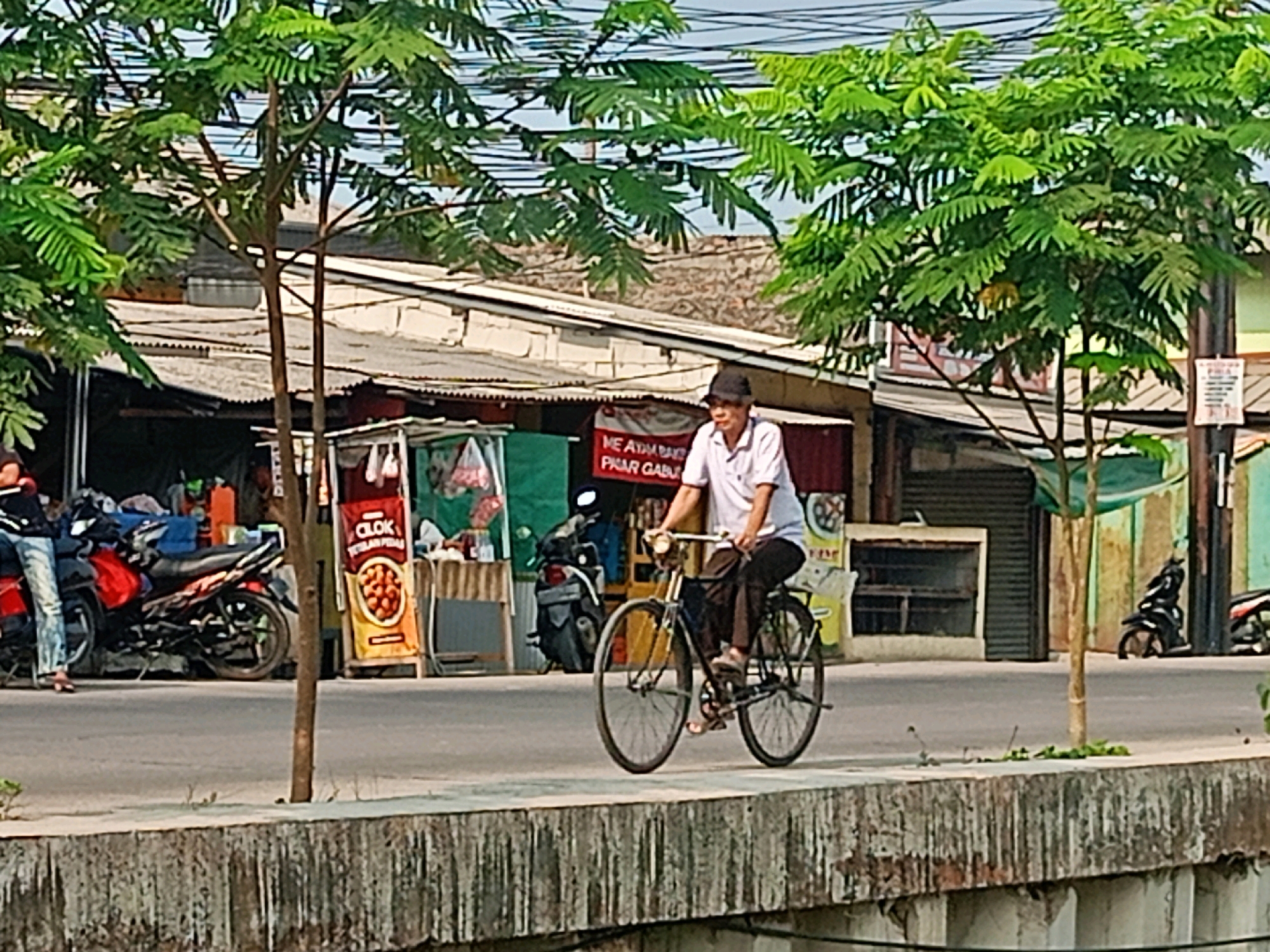 An elderly Indonesian man, wearing a white short-sleeved shirt and dark pants, rides a black bicycle on a paved road in Sriamur, Indonesia. He is also wearing a black hat. The man is the primary subject of the image and is captured in motion, with his right foot pushing down on the pedal. The bicycle is a vintage model with thin tires and a visible chain. In the background, there is a market stall with various items for sale, including what appears to be fried food or meatballs. A red motorcycle is parked to the left of the stall, and another motorcycle is visible further back. Buildings with tin roofs are situated behind the market. Lush green trees with dense foliage frame the scene from the left and right, with their branches extending across the top of the image. A concrete barrier runs along the bottom of the frame, separating the road from what appears to be a canal or body of water. The sky is overcast, suggesting a humid or cloudy day.