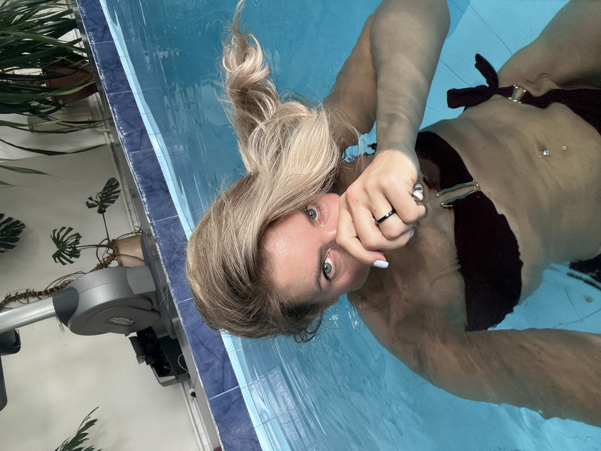 A woman is submerged in a swimming pool, with her upper body and face visible above the water. She has blonde hair, which is wet and splayed out around her head. She is wearing a dark-colored bikini top. One hand is raised to her face, covering her mouth and nose, and she is wearing a ring on her finger. The swimming pool has blue tiled walls. In the background, there is exercise equipment, specifically a stationary bike, and several potted plants. The scene appears to be indoors, possibly at a home or private gym facility, with a light-colored wall and ceiling visible. The lighting suggests it is daytime. The location is identified as Armavir, Russia.