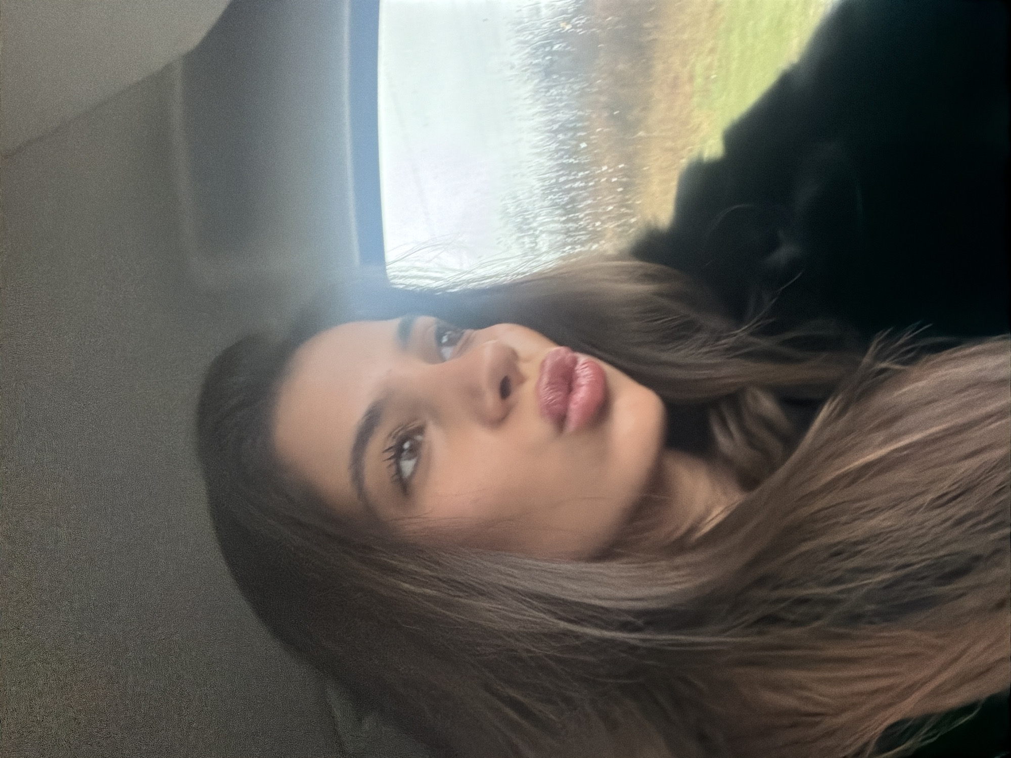 The image shows a woman with long brown hair and light skin. She has brown eyes and is puckering her lips. She is wearing dark clothing. She appears to be inside a vehicle, looking out the window. Raindrops are visible on the window. The background outside the window is blurry, but some green vegetation is visible. It is not possible to confirm the location as Dragichevo, Bulgaria, or provide additional context using only the content of the image.