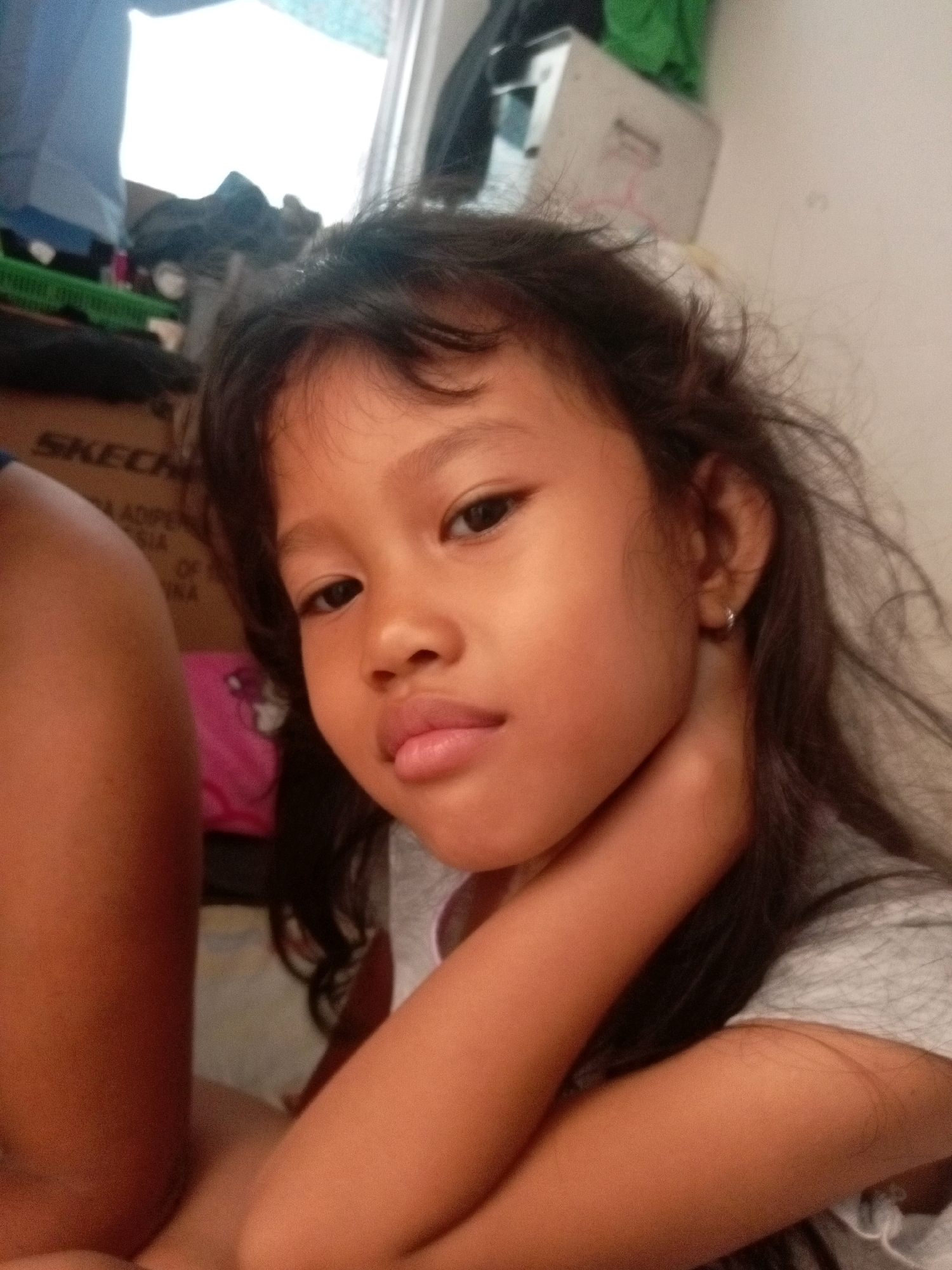 A young girl is posing for a selfie in what appears to be an indoor setting, possibly a bedroom. She has dark hair and is looking directly at the camera with a neutral expression. Her right arm is bent and resting on her chest, with her hand touching her neck. She is wearing a grey t-shirt. To her left, another person's arm, which appears to be darker-skinned, is visible.
In the background, there are some objects that suggest a living space. To the far left, a blue curtain or garment is partially visible. Below that, a green basket and what looks like a box with the word "SKECHERS" printed on it are present. There is also a pink object with a cartoon character on it behind the girl. The overall lighting suggests it could be daytime, with light coming from a window or bright light source behind the girl. The girl is wearing a small silver hoop earring in her visible ear. The scene has a casual and personal feel, typical of a home environment.