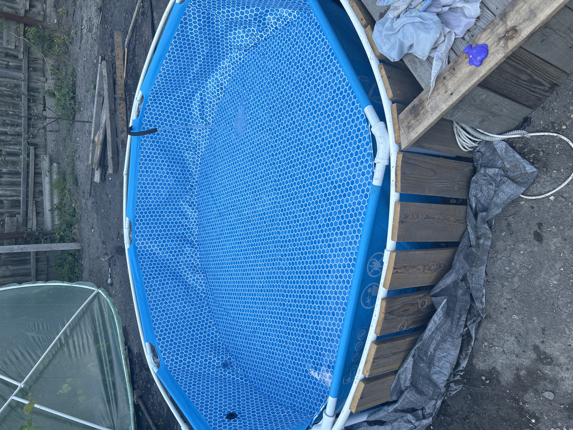 This is an eye-level, outdoor, full shot of a blue, hexagonal-patterned kiddie pool. The pool is empty and positioned on a dirt and gravel surface. To the right of the pool, there are several wooden planks, some of which appear to be part of a makeshift deck or platform. A purple plastic toy, possibly a frog, rests on one of the planks, next to a pile of white fabric. A white coiled hose is also visible near the wooden planks, and a dark grey tarp lies on the ground to the right, partially covering some of the planks. In the background to the left, a wooden fence and some greenery can be seen. Further left and slightly in the foreground, a portion of a green, geodesic dome structure, likely a greenhouse, is visible. The lighting suggests it is daytime, with overcast or diffused sunlight, indicating a cloudy or perhaps late afternoon sky. The scene appears to be in a backyard or a rural setting in Prokhladny, Russia. There is no visible text that can be clearly read.