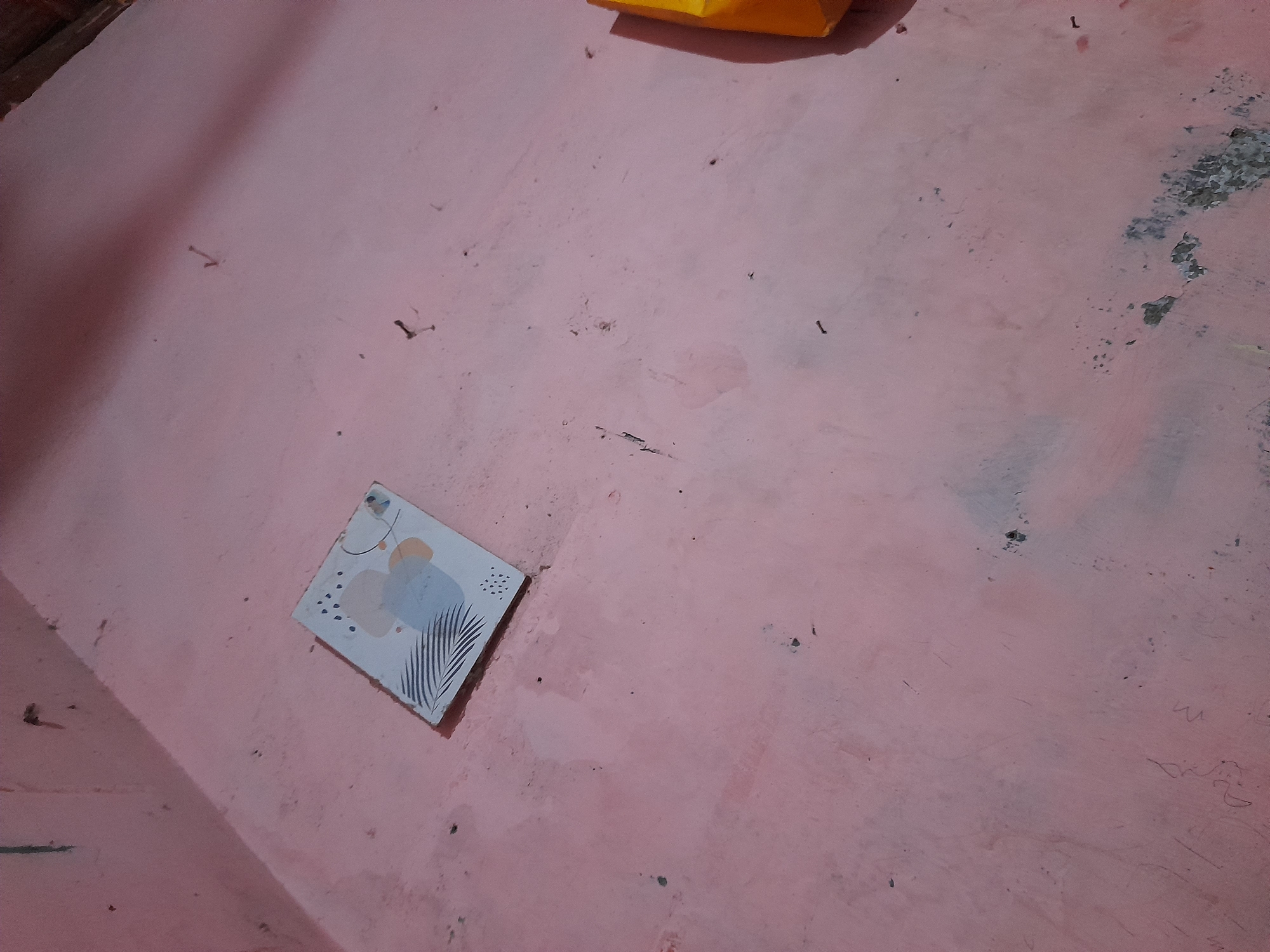 The image shows a section of a pink wall in what appears to be an indoor setting, possibly a room or hallway. A small, rectangular framed picture with an abstract design is mounted on the wall in the lower left quadrant. The design within the frame includes a light blue shape, an orange oval, and black lines resembling palm fronds. The wall itself is textured and shows signs of age and wear, with some darker marks and smudges scattered across its surface. The lighting suggests it might be daytime, with a strong light source casting a shadow diagonally across the left side of the frame, indicating a light source from the upper left. There are several small, dark objects that resemble nails or tacks protruding from the wall in various locations. In the upper right portion of the image, a sliver of a yellow object, possibly a bag or piece of fabric, is visible. The overall impression is of a simple, somewhat worn interior space. No people are visible, and no discernible text is present.