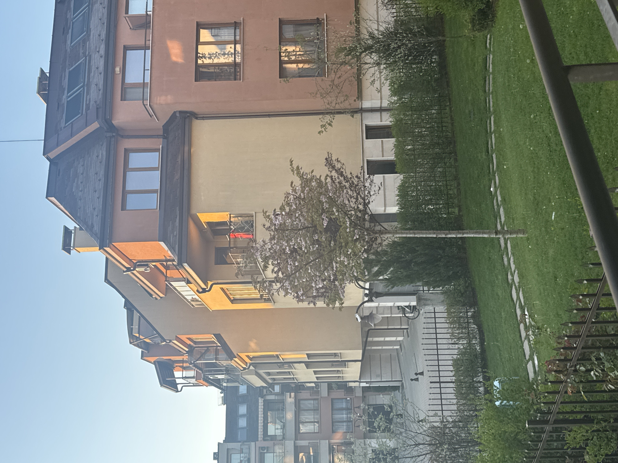 The image depicts a residential area in Sofia, Bulgaria, characterized by modern apartment buildings and a landscaped courtyard. The buildings are multi-story structures with a variety of architectural styles and facade colors ranging from brown and orange to yellow and grey. Each building features numerous windows, some with visible reflections. A narrow green courtyard separates the buildings, featuring grass, hedges, and a flowering tree in the center. A pathway made of stone slabs cuts through the grass. Fences run along the perimeter. The sky is clear and blue, suggesting a sunny day. The image is taken from an elevated perspective, possibly from a window or balcony. No people are visible.