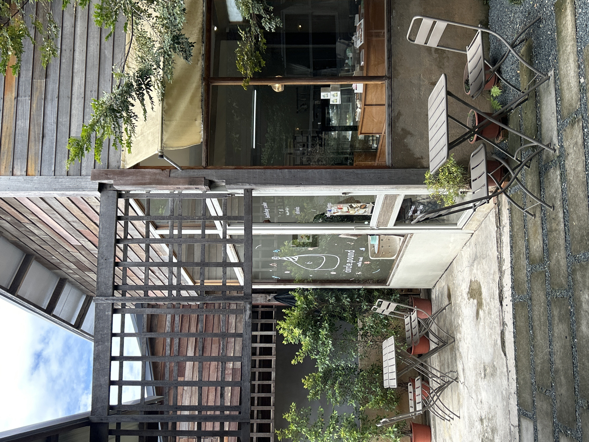 An exterior, eye-level, medium shot captures a quaint cafe's seating area situated in Chiang Mai City Municipality, Thailand. Two small, grey metal bistro tables, each accompanied by two matching chairs, are arranged on a textured concrete patio. The tables are set with a simple, clean aesthetic. Behind the tables and chairs, lush green bushes and potted plants add a touch of nature to the scene. A large windowpane of the cafe is visible to the right, revealing a glimpse of the interior with display cases showcasing pastries and baked goods. The cafe's architecture features wooden siding and a dark wooden lattice structure, contributing to its rustic charm. The sky above is partly cloudy, indicating fair weather. The overall impression is one of a peaceful and inviting outdoor dining space within an urban setting.