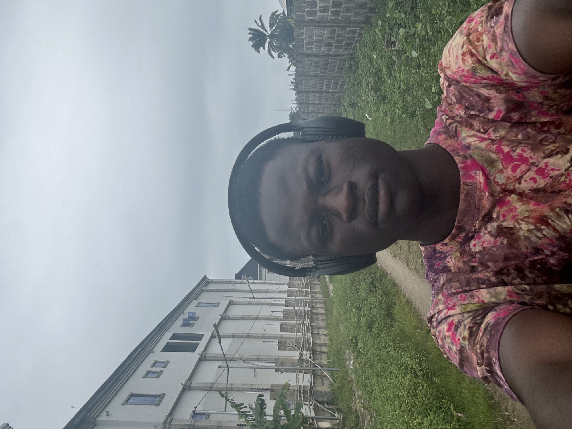 The image is a selfie taken outdoors in Badagry, Nigeria, under an overcast sky. The subject, a young Black male wearing black headphones and a t-shirt with a pink, purple, and green tie-dye pattern, is positioned in the foreground, facing the camera. Behind him, to the left, is a large, multi-story building with a white facade, featuring numerous windows and columns. A fence made of concrete blocks and wooden posts, with some wire fencing, runs along the front of the building. To the right of the subject, a brick wall extends into the background, with a palm tree visible above it. The ground appears to be a dirt path with overgrown green vegetation on either side.