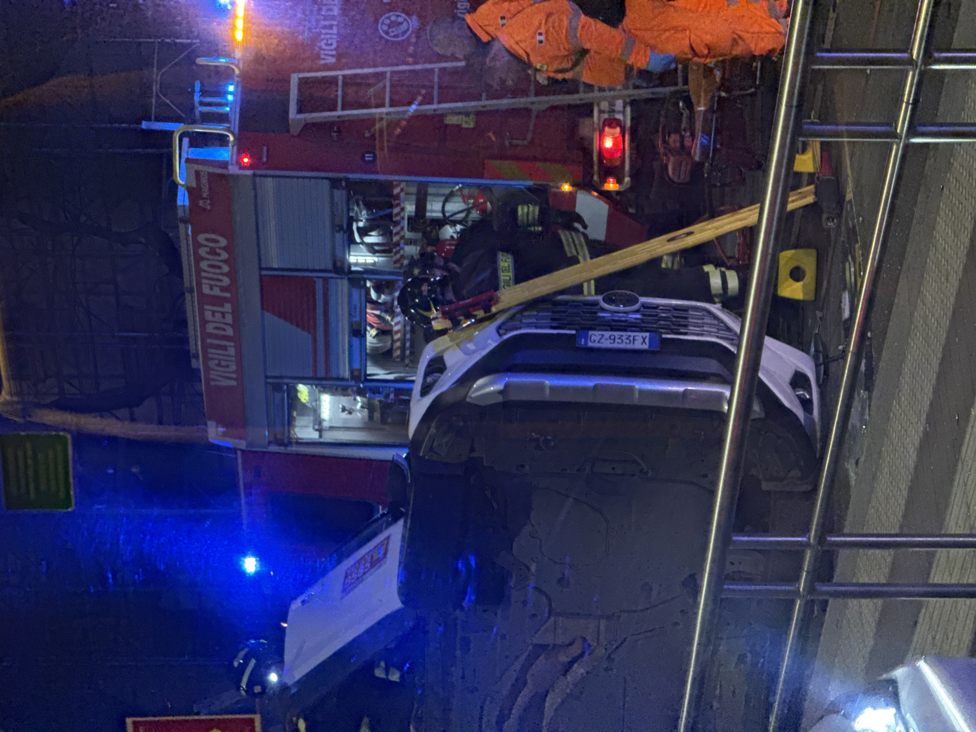 An urban nighttime scene in the city of Milan, Italy, depicts an emergency response to an overturned white SUV. The SUV rests on its left side, with its undercarriage and front grille facing the viewer. An Italian license plate, "EZ-933FX," is visible on the front of the vehicle. A red "VIGILI DEL FUOCO" (Fire Brigade) Magirus fire truck is positioned behind the SUV, its blue and orange emergency lights illuminating the area. A firefighter in a dark uniform and helmet is near the SUV, and another individual in a bright orange rescue suit is observed near the rear of the fire truck. A long wooden plank or ladder leans against the overturned vehicle. A silver metal railing runs across the foreground, partially obscuring the lower portion of the scene. The ground is paved, with minor debris visible.