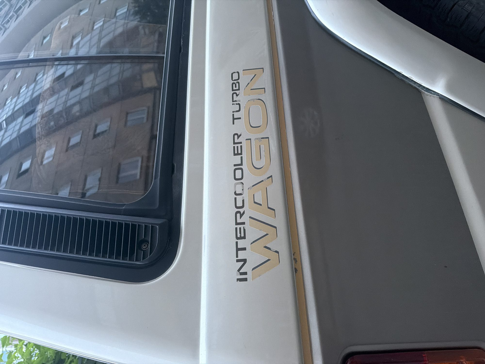 The image shows a close-up of the side of a white and gray vehicle, with a gold decal that reads "INTERCOOLER TURBO WAGON." The vehicle appears to be parked outdoors, with a tall building reflected in the window. The building is modern, with many windows, and the reflection suggests it is daytime. The ground is not visible in the image. The scene is a close-up of a vehicle's branding.