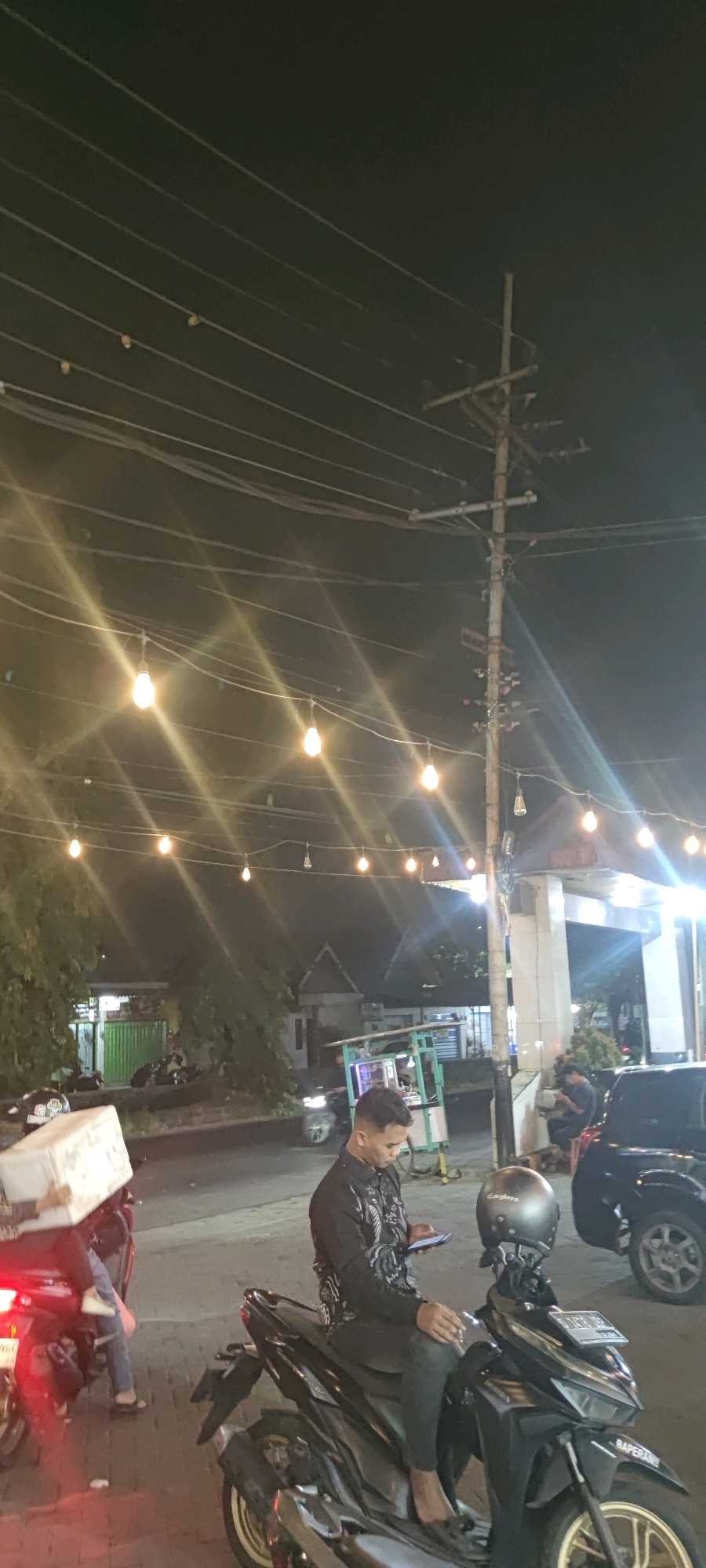 The image captures a street scene at night in Keboansikep, Indonesia, illuminated by strings of decorative lights and ambient street lighting. In the foreground, a person wearing a black patterned shirt and dark pants is seated on a black motorcycle, looking down at a mobile phone. A silver helmet rests on the handlebars of the motorcycle. To the left, another motorcycle is partially visible, carrying a large white box on its rear. In the background, trees and buildings are dimly lit. Power lines and a utility pole are prominent elements in the sky, with several hanging light bulbs extending across the scene. A black vehicle is parked to the right of the frame.