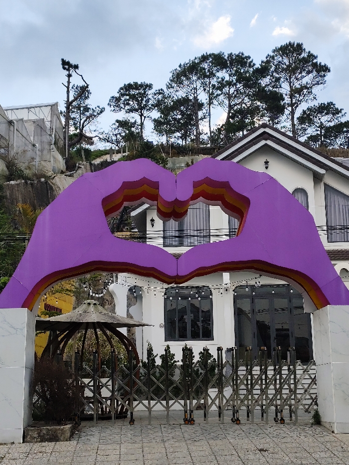 The image captures an exterior view in Thành phố Đà Lạt, Vietnam, centered on a large, purple archway designed as two hands forming a heart. The archway features interior concentric bands of orange, yellow, and red. It is situated on a paved surface composed of light-colored interlocking tiles.
Behind the archway, a two-story white building with a brown tile roof and prominent dark-framed windows is visible. Some windows display grey curtains, and multiple exterior wall sconces are affixed to the facade. A fully extended, accordion-style stainless steel gate blocks access to the building's entrance. Overhead string lights are strung across the building's exterior, positioned above the gate. To the left of the gate, a weathered, round, umbrella-like structure, possibly a gazebo roof, is partially visible, alongside a desiccated plant in a dark planter.
The background features a steep hillside densely covered with numerous mature pine trees. A white-framed greenhouse structure is observable on the upper left slope. The sky is largely overcast. No human figures are present within the frame.