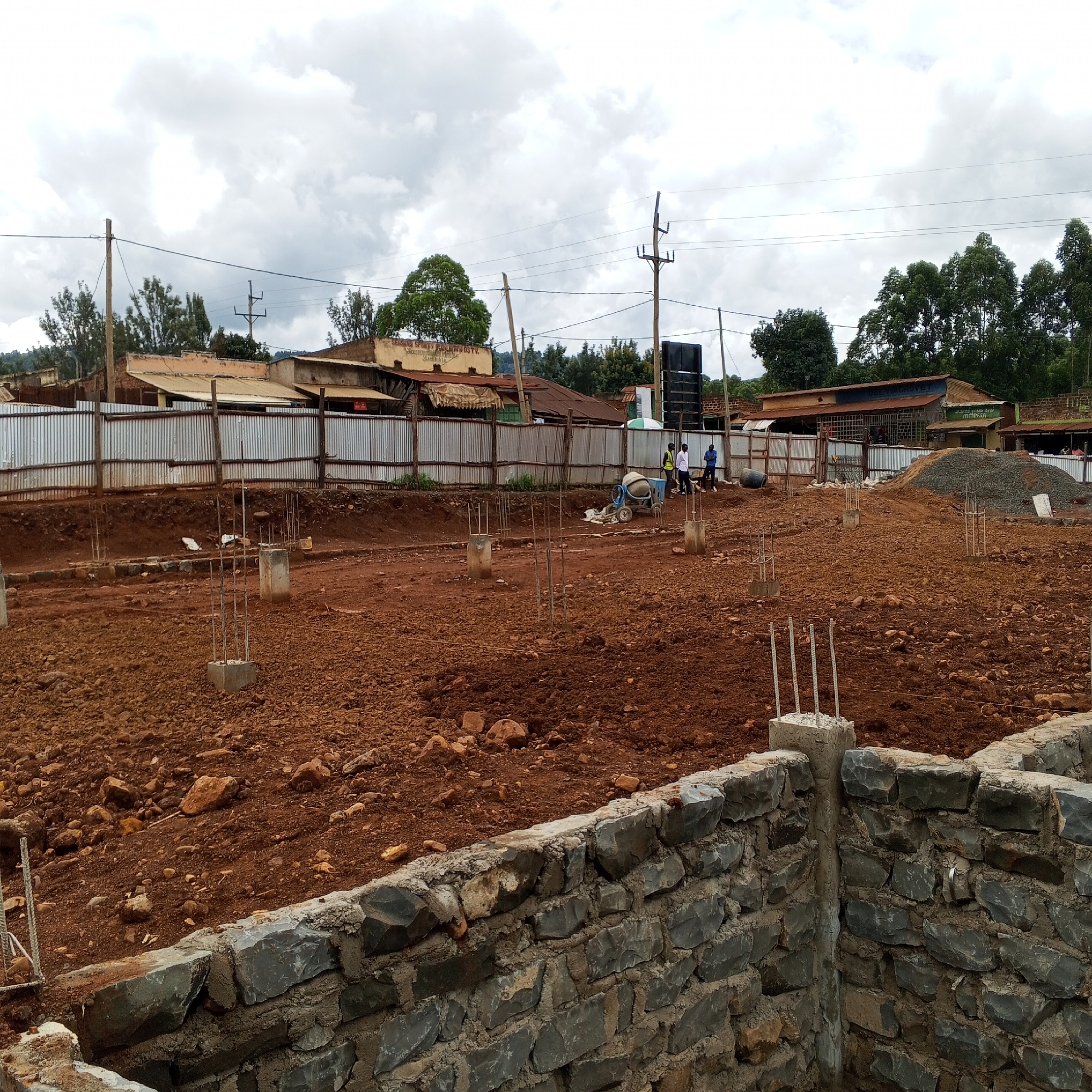 The image captures an active construction site in Borabu Chitago ward, Kenya, under a cloudy, overcast sky during the daytime. In the immediate foreground, a robust stone foundation wall, constructed from dark, irregularly shaped stones, is partially built. Beyond this, the ground is a expanse of reddish-brown soil, indicative of recent excavation, dotted with rocks and partially completed concrete pillars with rebar extending upwards, marking the future structure's footprint.
In the mid-ground, construction activity is underway with a blue concrete mixer visible. Several workers are present; three men are standing near the mixer, with one appearing to operate it, while another worker stands further to the right. Behind the construction area, a corrugated metal fence separates the site from the adjacent properties.
The background reveals a local commercial or residential area with various buildings. These structures generally feature corrugated iron roofs and simple facades. Visible text on a building to the left reads "SEASONS SPECIAL QUALITY HOSTEL." Further to the right, another building displays a sign for "ALLE AGROVET" and a smaller "m-pesa" sign beside it. Wooden utility poles with power lines stretch across the frame, connecting to the nearby buildings. Lush green trees are scattered throughout the background, adding to the semi-urban landscape. The scene conveys a sense of ongoing development and community life.