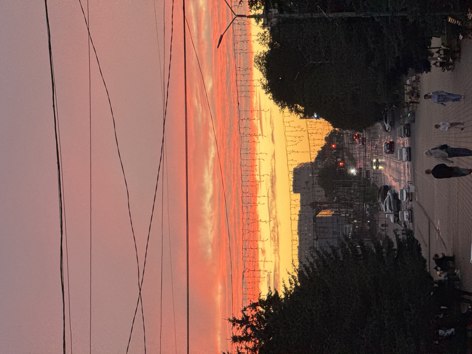 A vertical, medium-shot photograph captures a sunset over a city street in Mikhaylovsk, Russia. The sky displays vibrant hues of pink, orange, and yellow. Overhead power lines crisscross the frame, and a string of lights hangs horizontally across the middle. Silhouetted trees frame the scene on the left and right. In the background, buildings line the street, and traffic, including cars and buses, moves along the road. Several individuals are visible on a paved walkway, some walking and others sitting on benches. The overall impression is of a tranquil evening scene in a city.