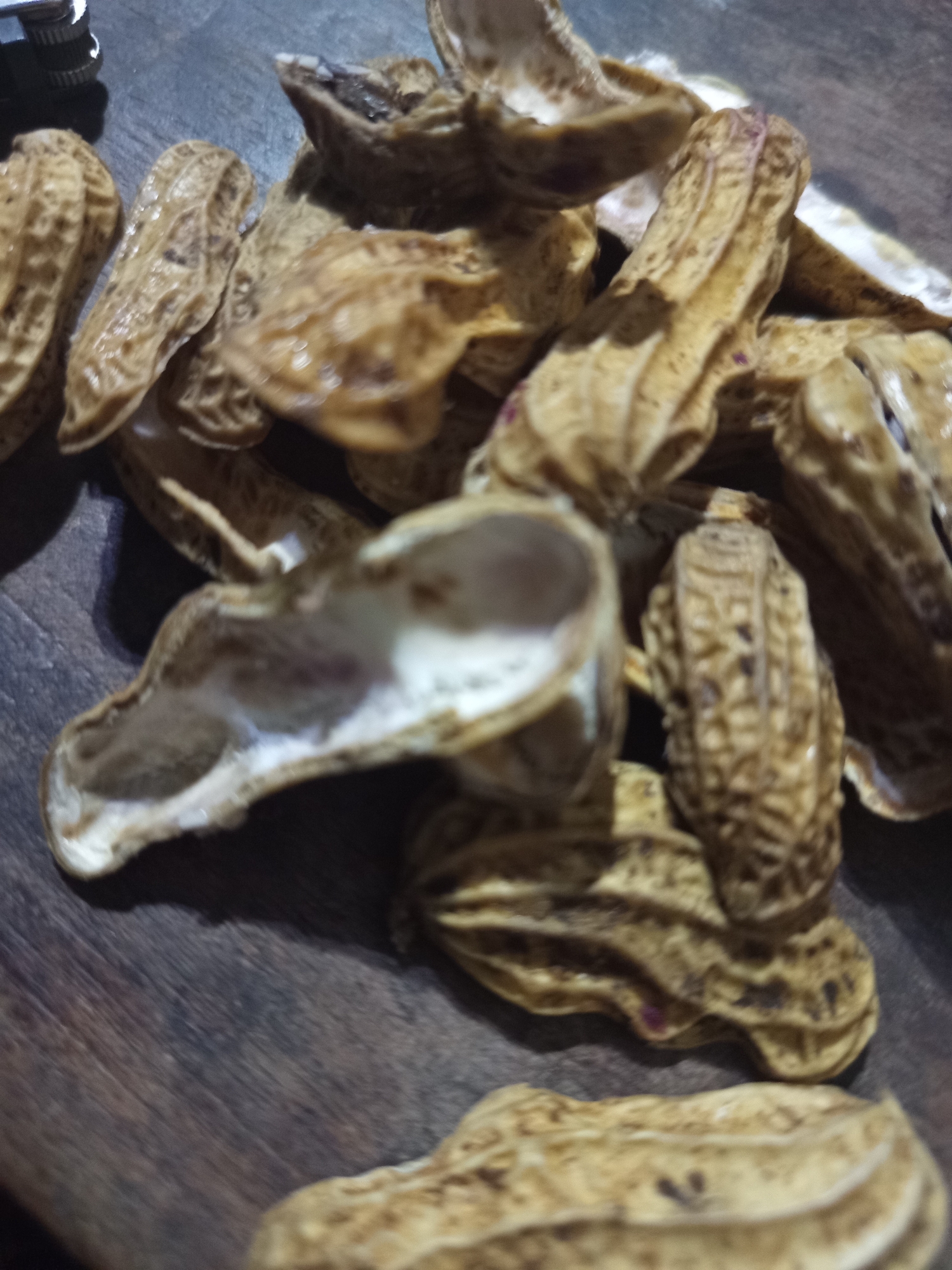 The image displays a pile of empty peanut shells on a dark, textured surface, possibly wood. The shells are light brown and appear to be dry, with some showing slight discoloration or remnants of the peanut. The arrangement is haphazard, suggesting they have been discarded. The lighting is dim, casting shadows that add to the overall somber tone. There are no people or any other identifiable objects in the scene. The image does not contain any contextual information that would indicate its location in Ngablak, Indonesia.
